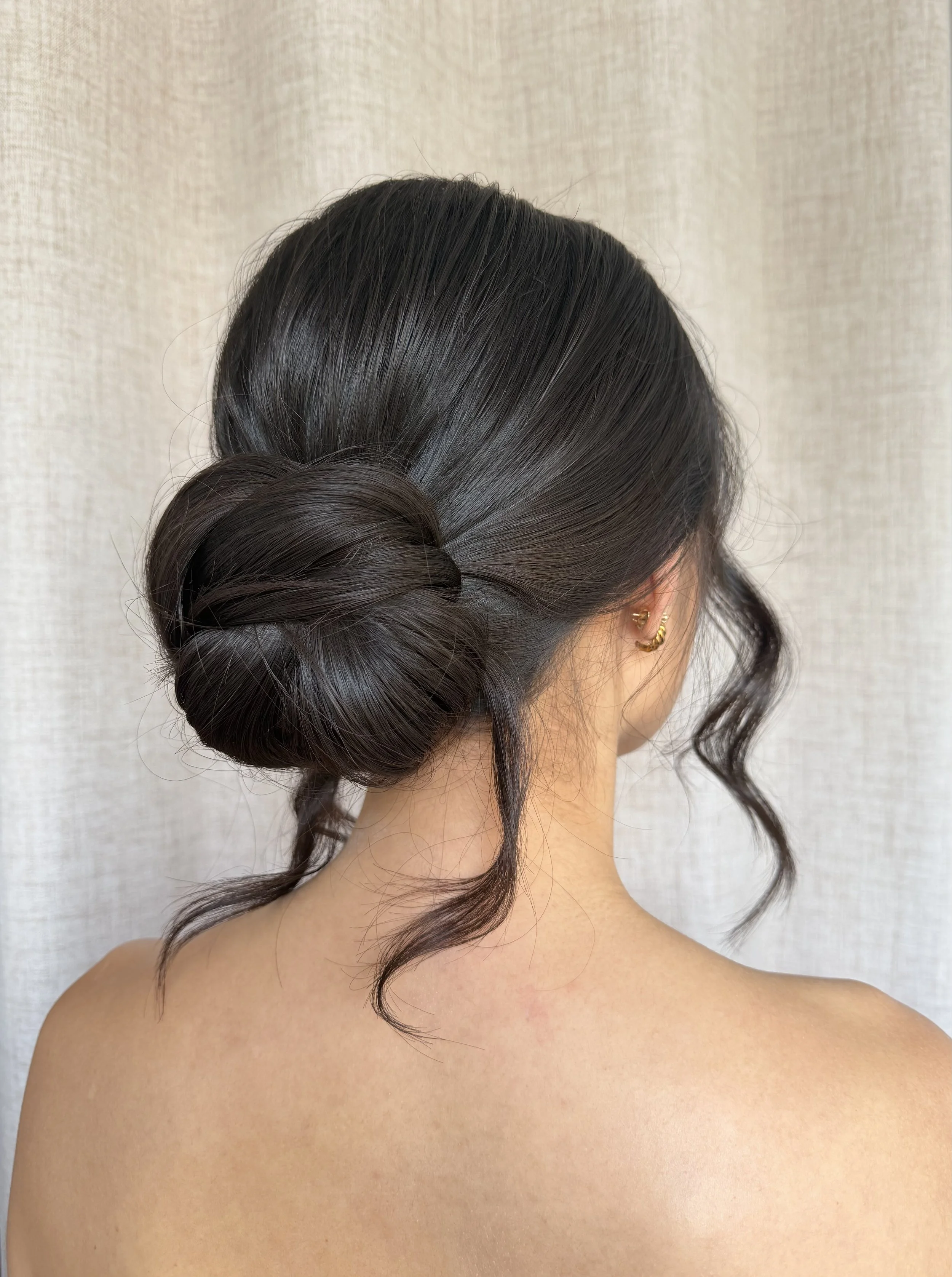 Back of a person's head with an elegant styled updo hairstyle and curls at the lower part of the hair, with visible hair color and a gold hoop earring, against a beige fabric background.