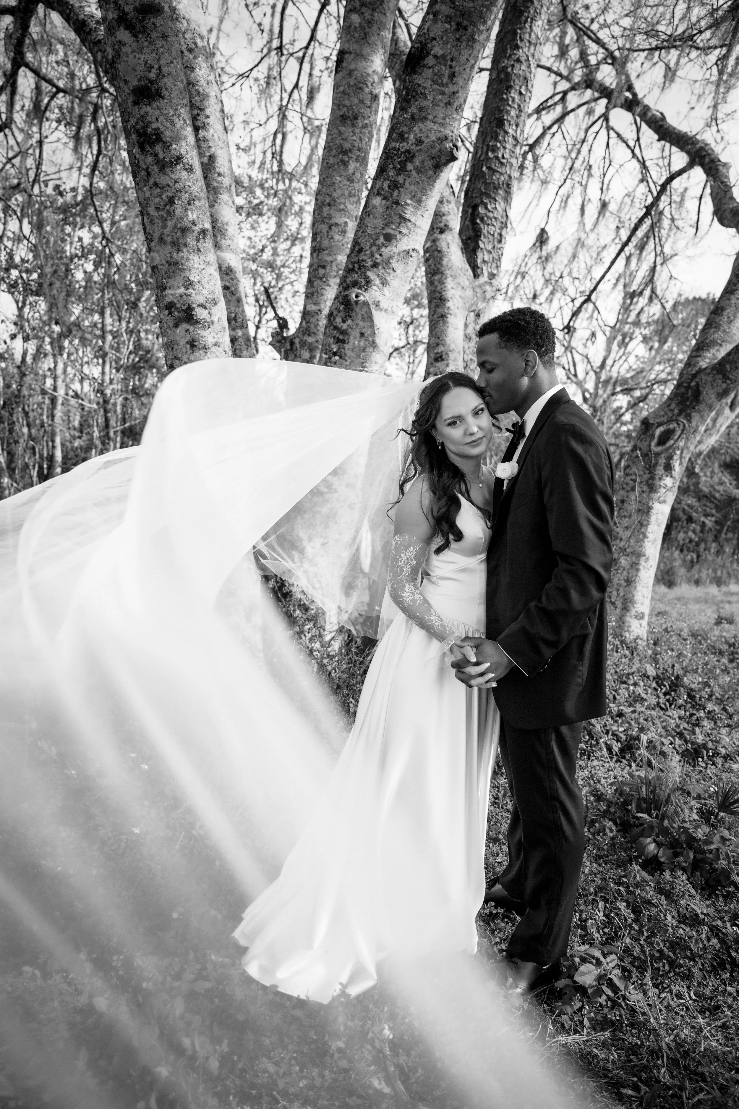 A black-and-white photograph of a bride and groom standing close together outdoors among trees. The bride is wearing a white gown and veil, and the groom is in a dark tuxedo. They are holding hands, and the groom is kissing the bride on her forehead while she looks at the camera. The scene has a natural, serene backdrop with leafless trees and a cloudy sky.