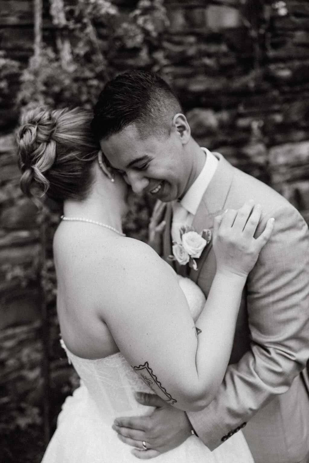 A black and white photo of a bride and groom sharing a close, joyful embrace on their wedding day, with a stone wall in the background.