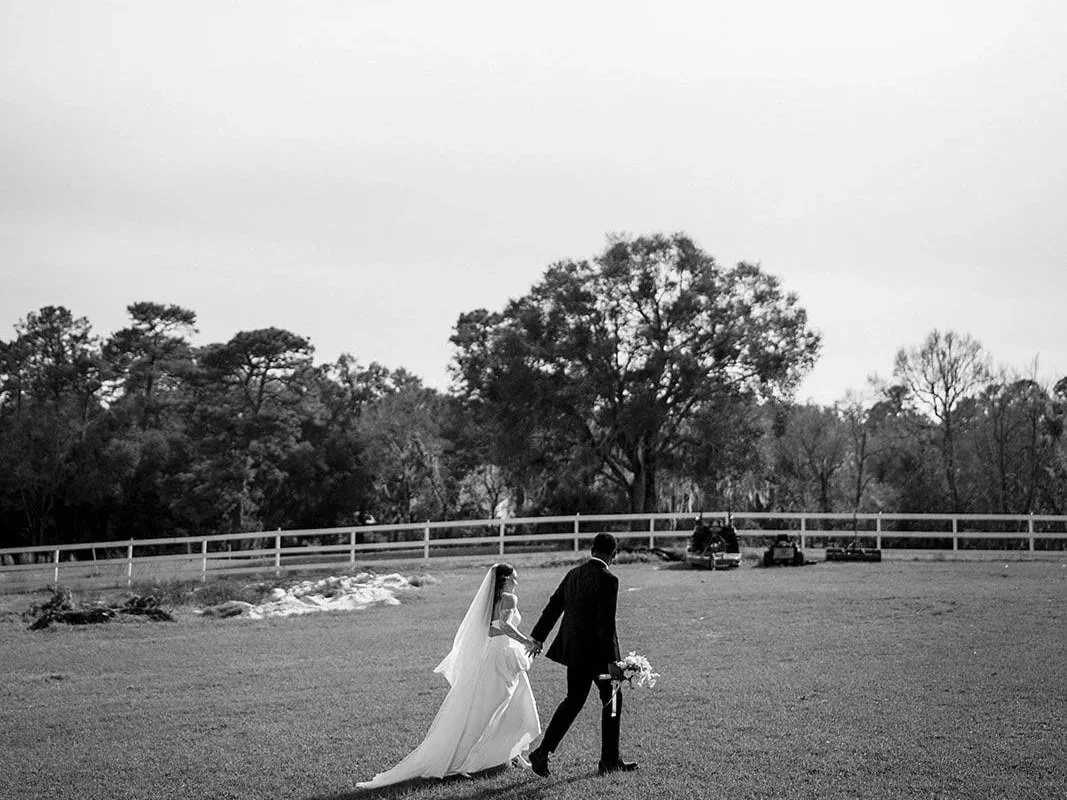 A wedding couple walking hand in hand across a grassy field with trees in the background, a white fence, and some vehicles parked nearby.