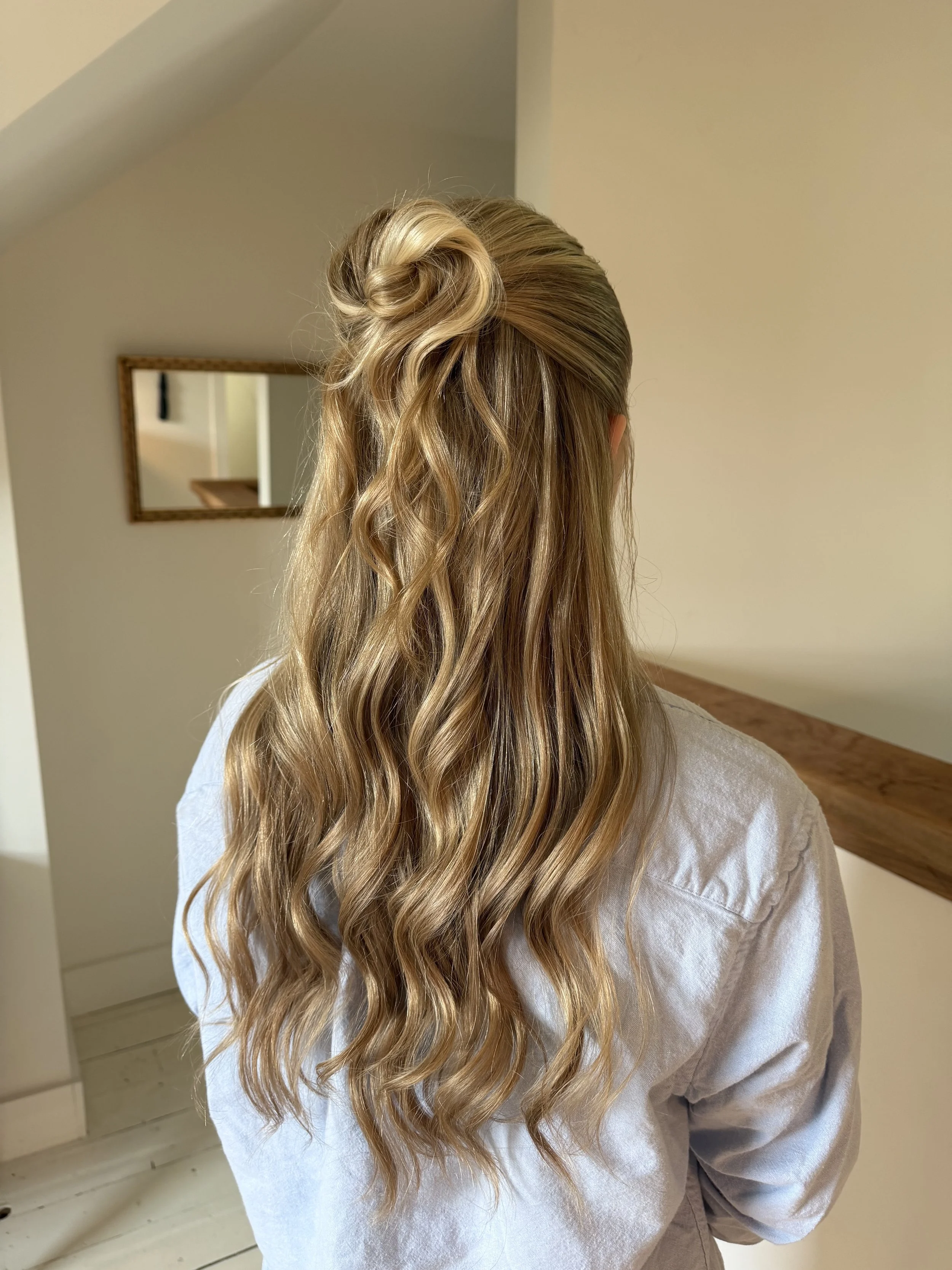 Back view of a woman with long, wavy blonde hair styled in a twisted half-updo, wearing a beige or light-colored shirt, standing in a room with a wall mirror and wooden furniture.