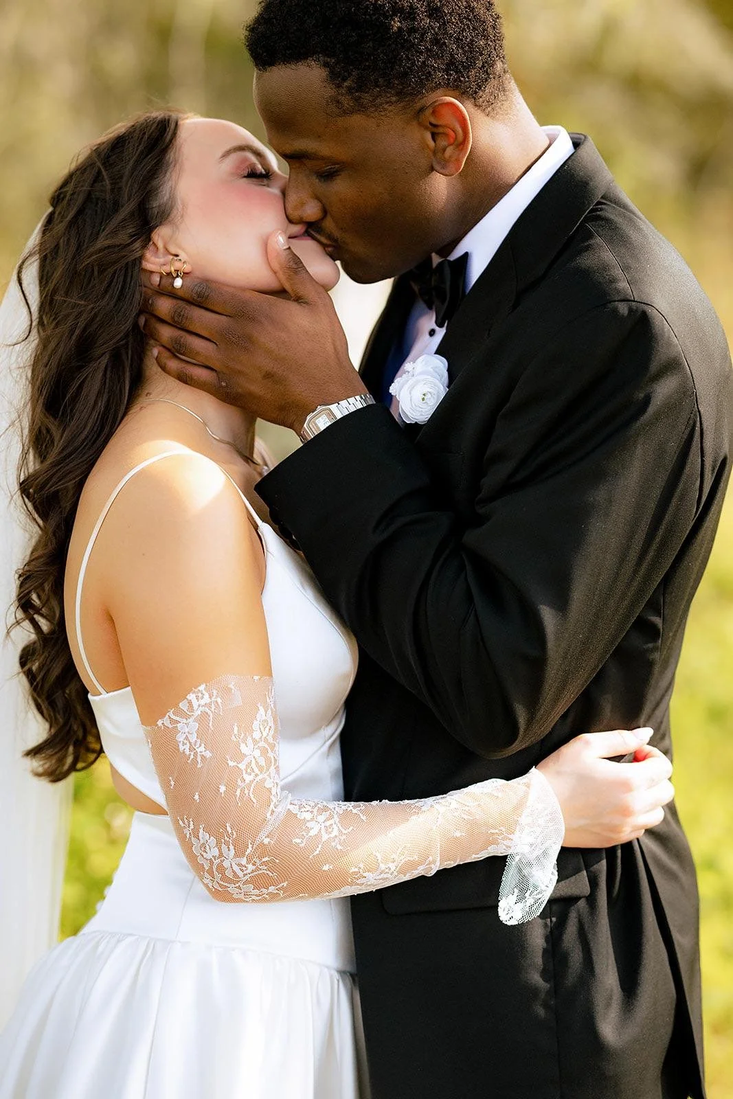 A bride and groom embrace and kiss outdoors, with the groom holding the bride's face, both dressed elegantly for their wedding.
