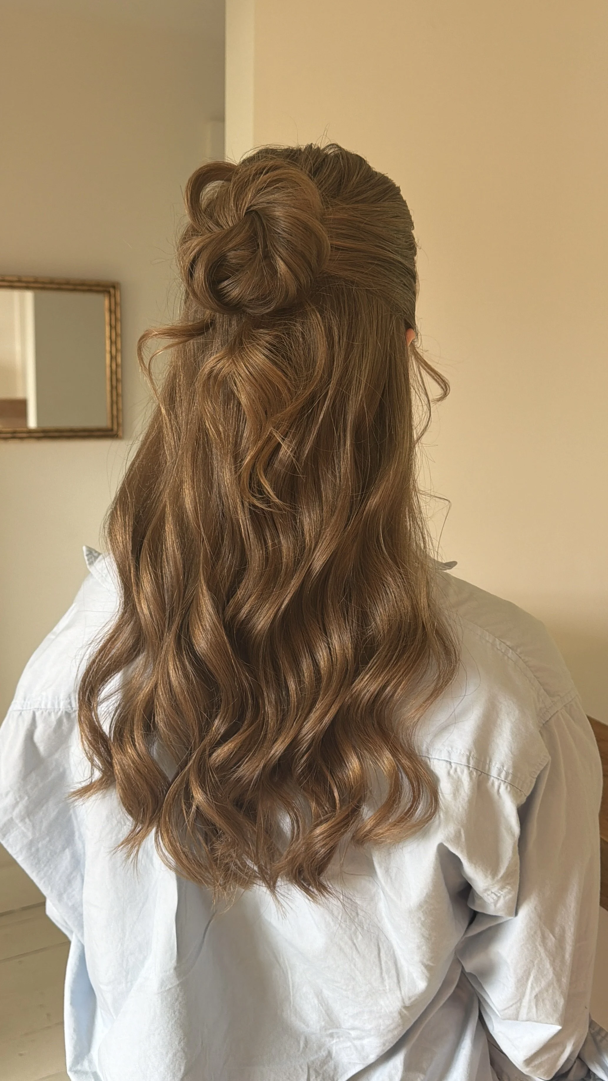 Back view of a woman with long, wavy, light brown hair styled in loose curls and an elegant twist at the crown, wearing a white shirt, in a room with beige walls and a mirror.