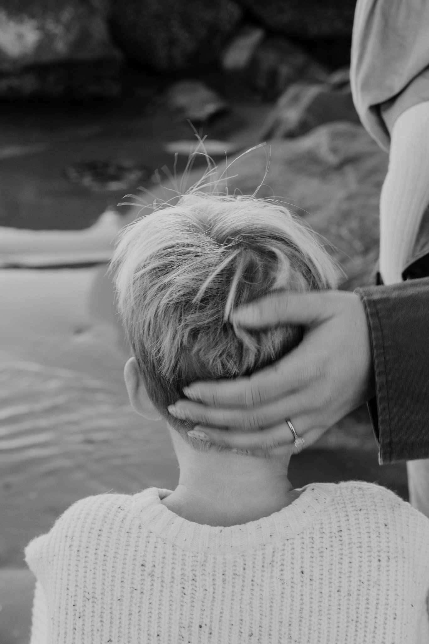 A person with short hair receiving a blessing from another person, with their hand gently placed on the back of the head by a third person. The scene appears to be outdoors near water.