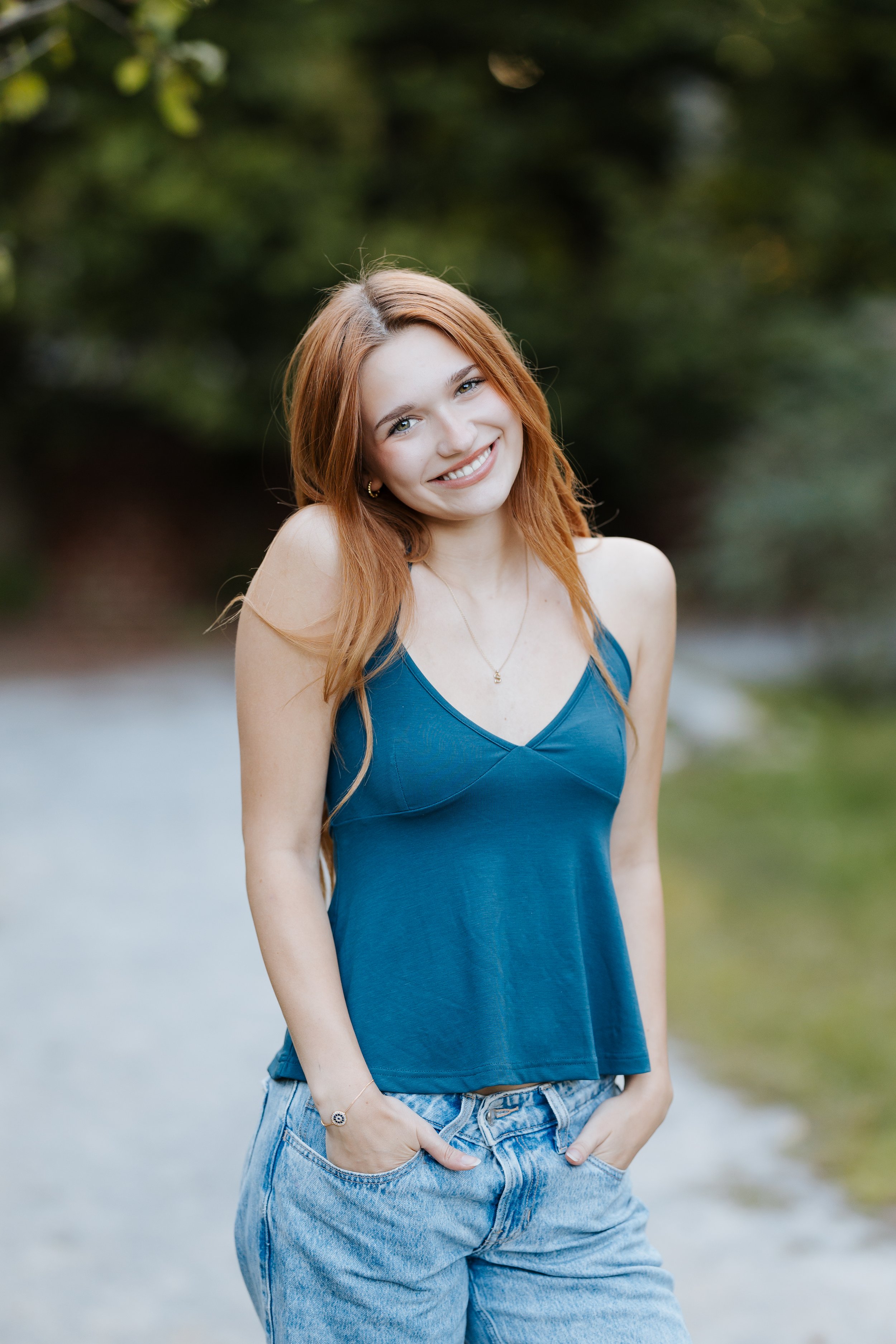 A young woman with red hair and fair skin smiling outdoors, wearing a teal tank top and light blue jeans.