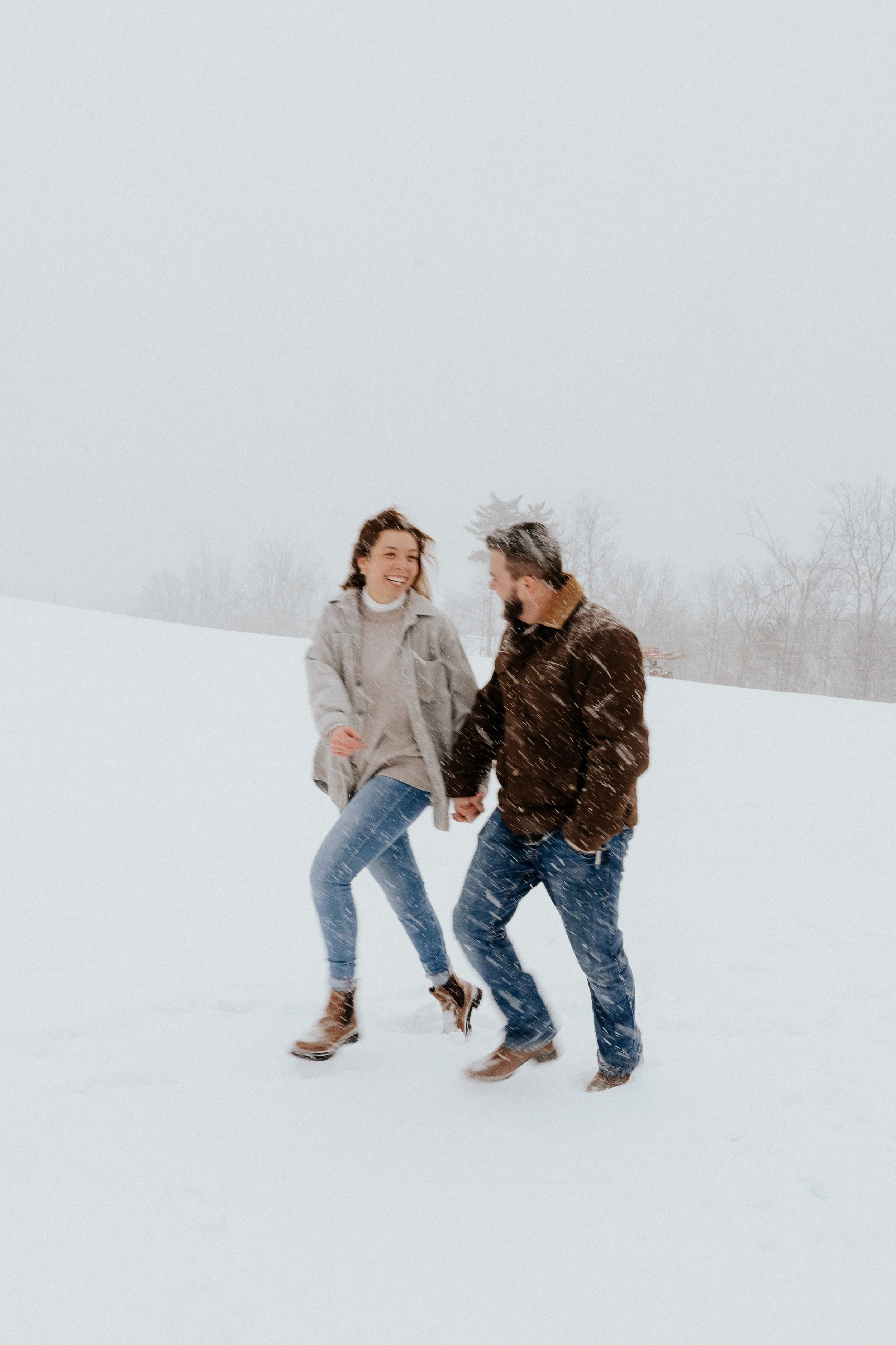 A couple holding hands and playing in the snow, smiling, with falling snow and leafless trees in the background.