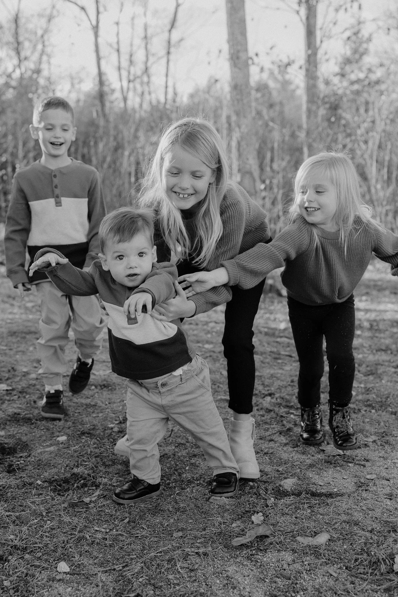 Four children, two girls and two boys, playing and laughing outdoors in a wooded area on a fall day.