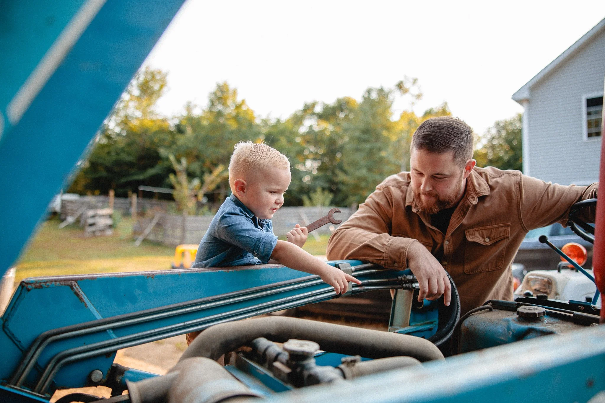 A young boy and an adult male working on a vehicle outdoors, with trees and a house in the background.