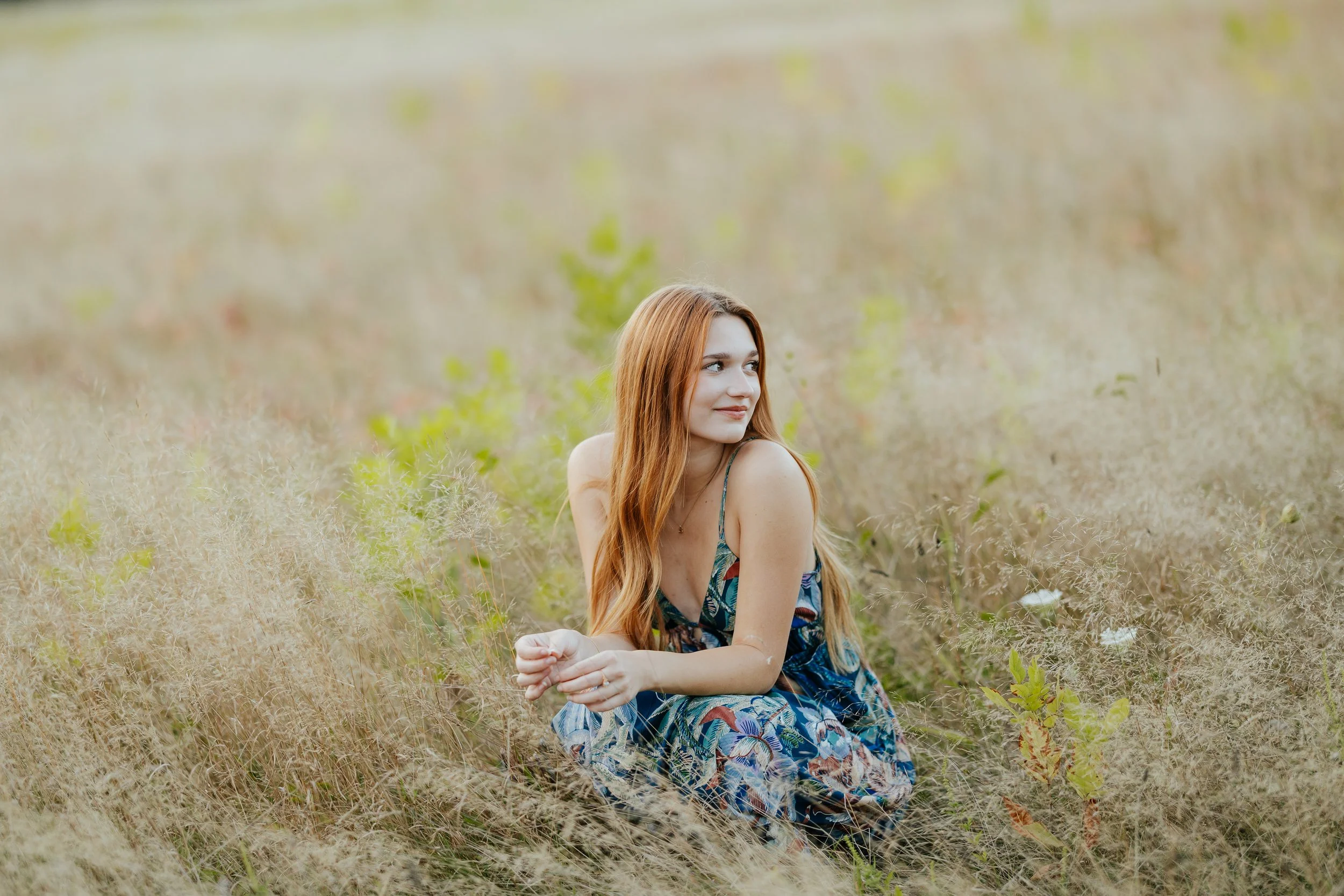 A young woman with long red hair sitting in a grassy field, wearing a colorful patterned dress, looking to her right with a gentle smile.