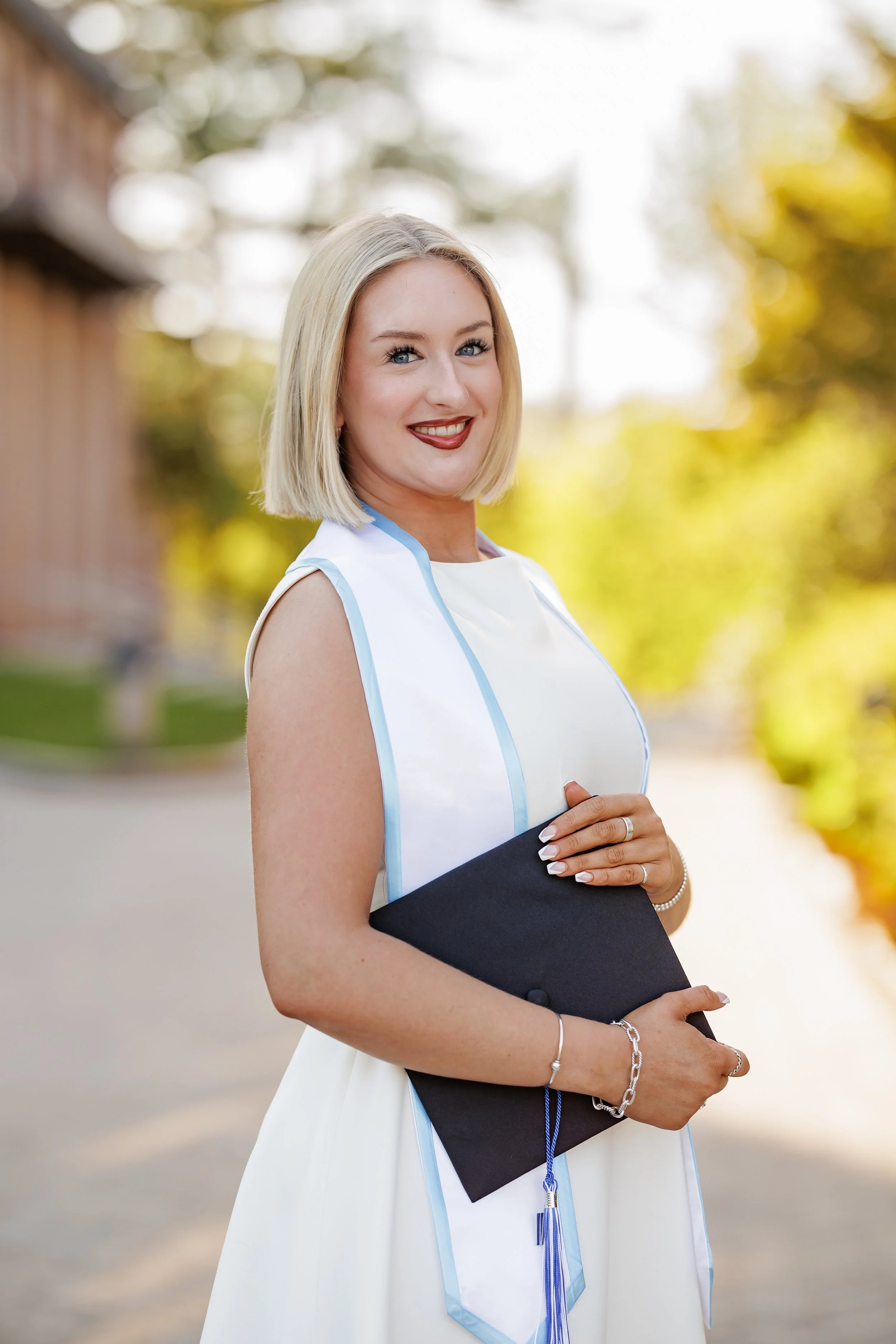 A young woman in a white graduation dress holding a black diploma folder outdoors with a blurred background of trees and a building.