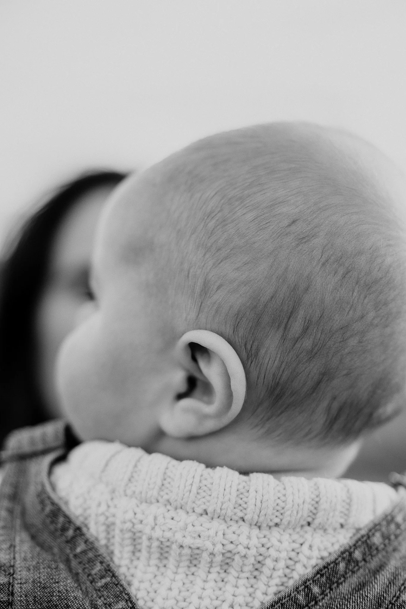 A black and white close-up photograph of a child's head showing the back and side with a shaved hairstyle, wearing a knitted sweater and denim jacket.