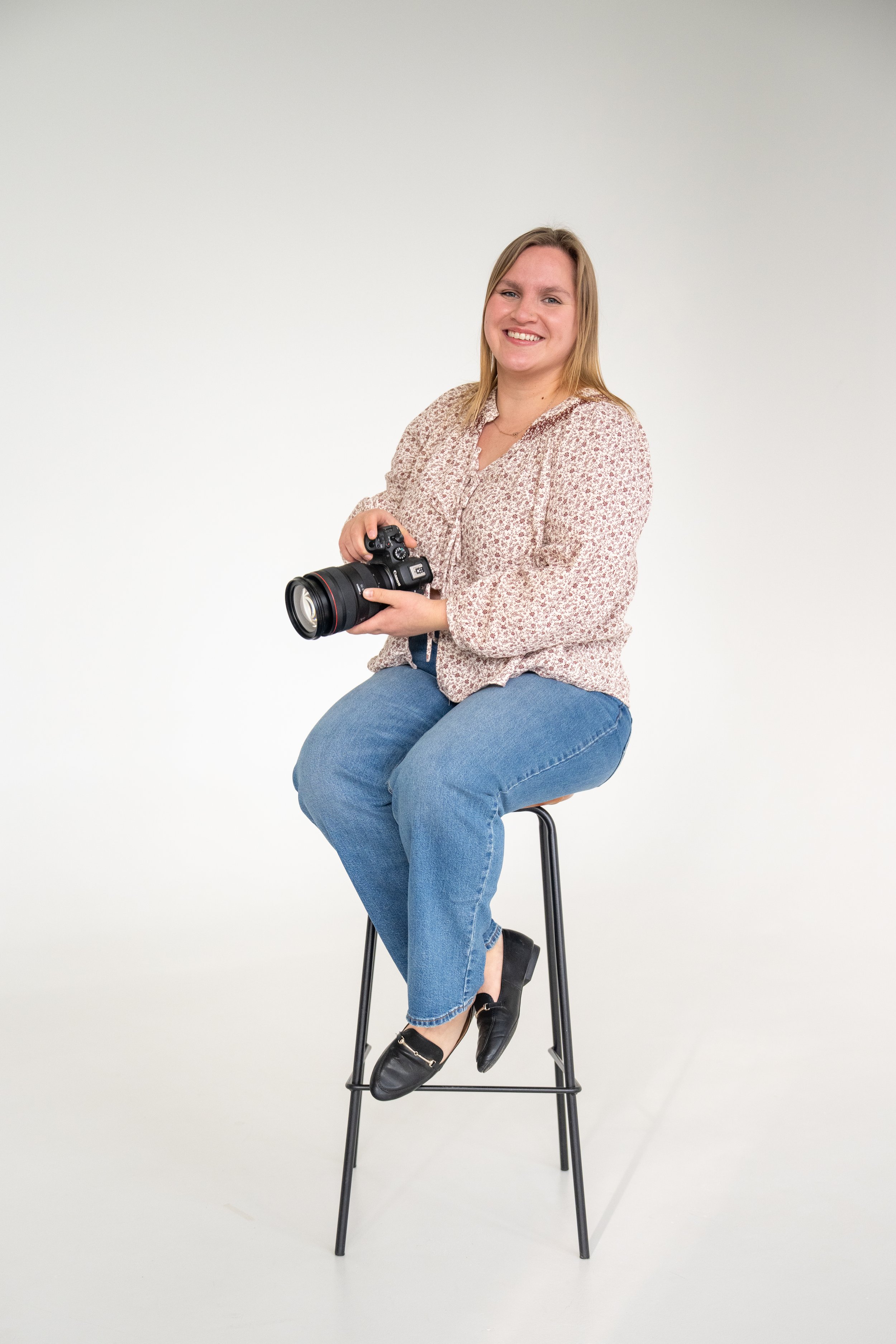A woman with blonde hair smiling, holding a camera, sitting on a tall stool against a white background.