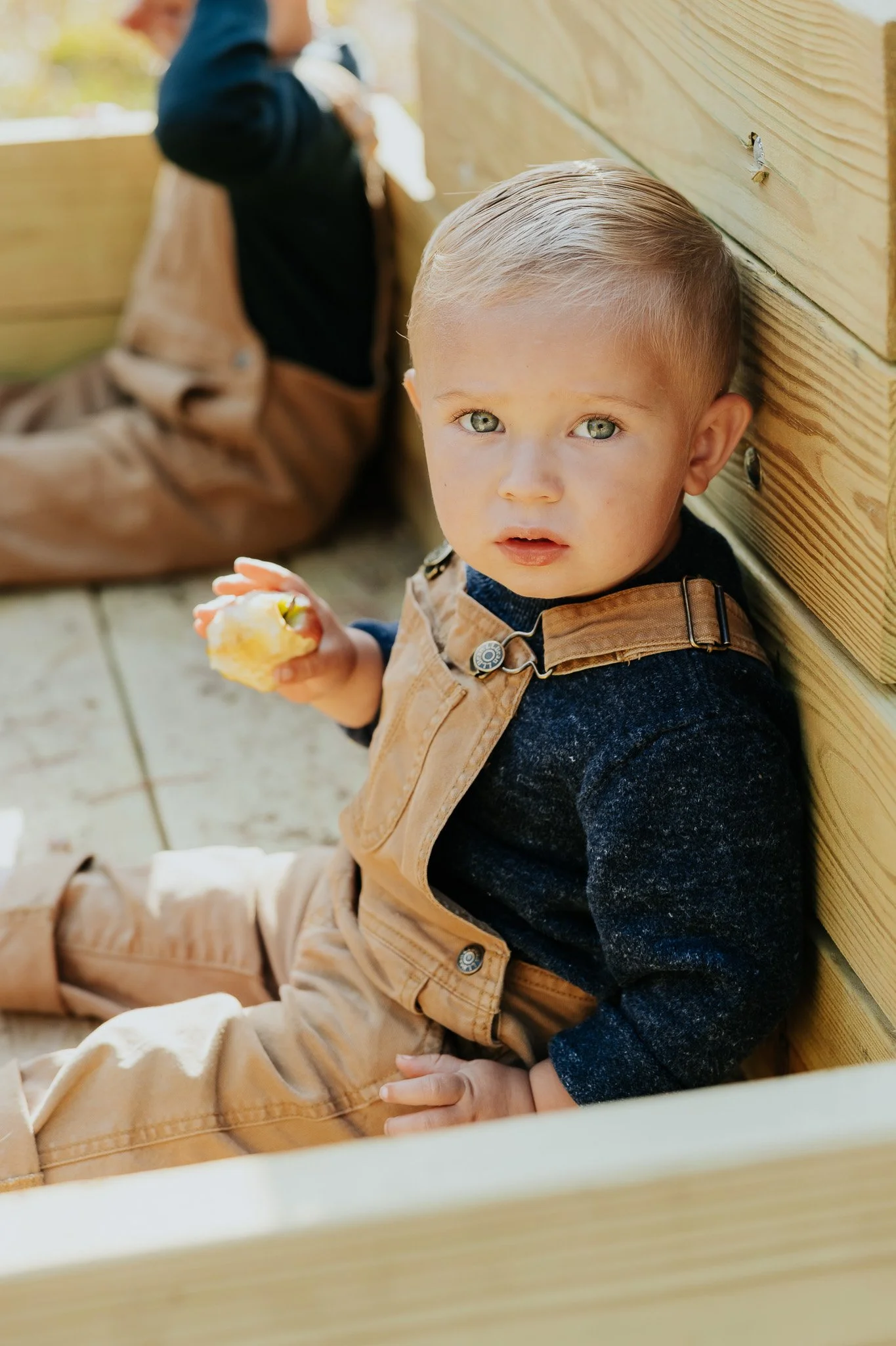 A young boy with blond hair and blue eyes sitting on a wooden deck, holding a partially eaten snack, and looking at the camera. He is wearing tan overalls over a dark long-sleeve shirt.