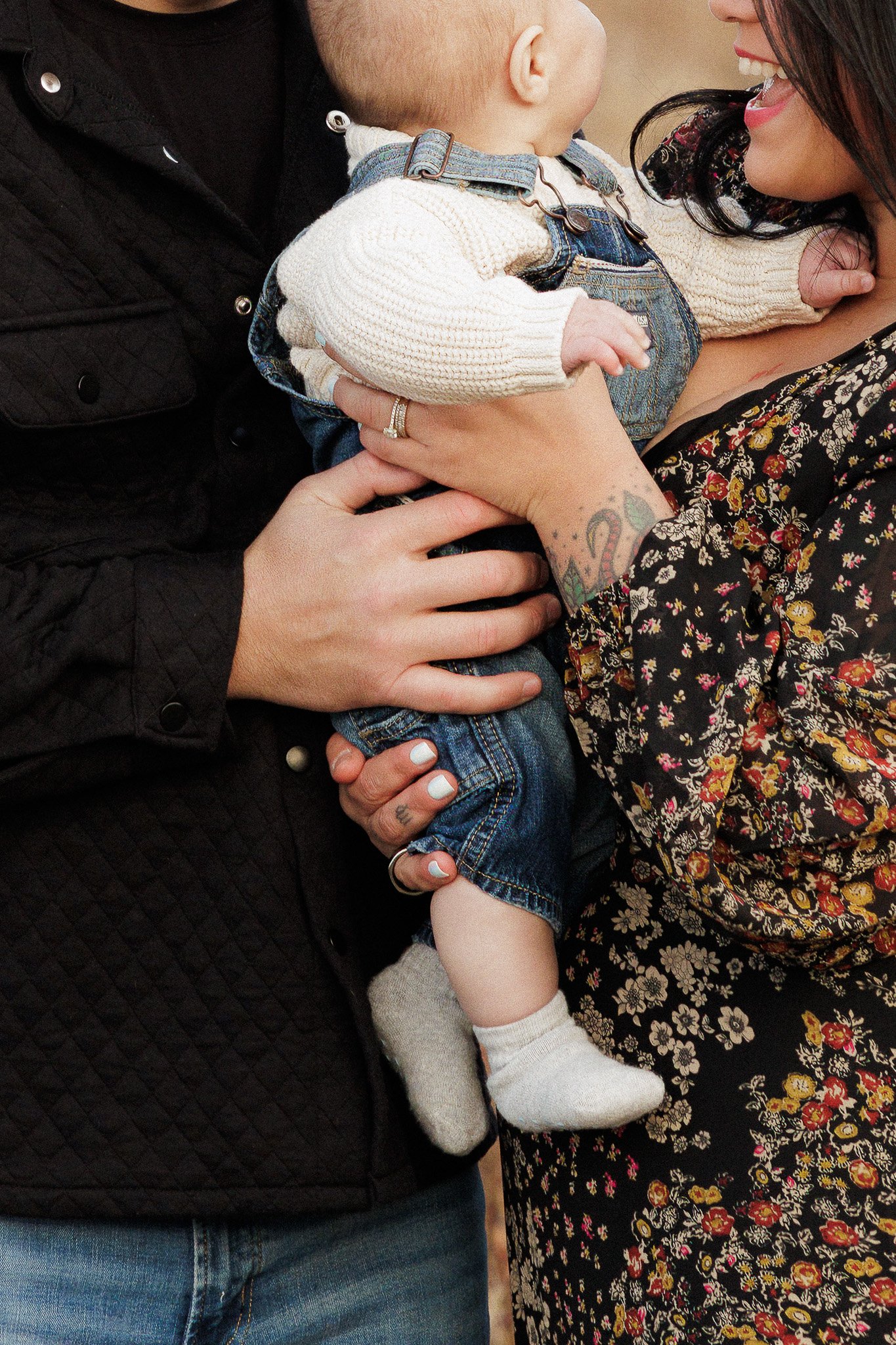 Close-up of a woman and man holding a baby, smiling and looking at each other. The woman has a tattoo on her arm and is wearing a floral dress. The man is wearing a black quilted jacket and blue jeans. The baby is dressed in overalls, a cream sweater