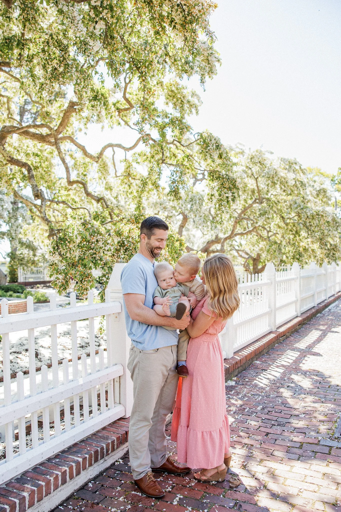 Family of four standing on a brick sidewalk near a white picket fence, under a large blooming tree, smiling and interacting with each other during daytime.