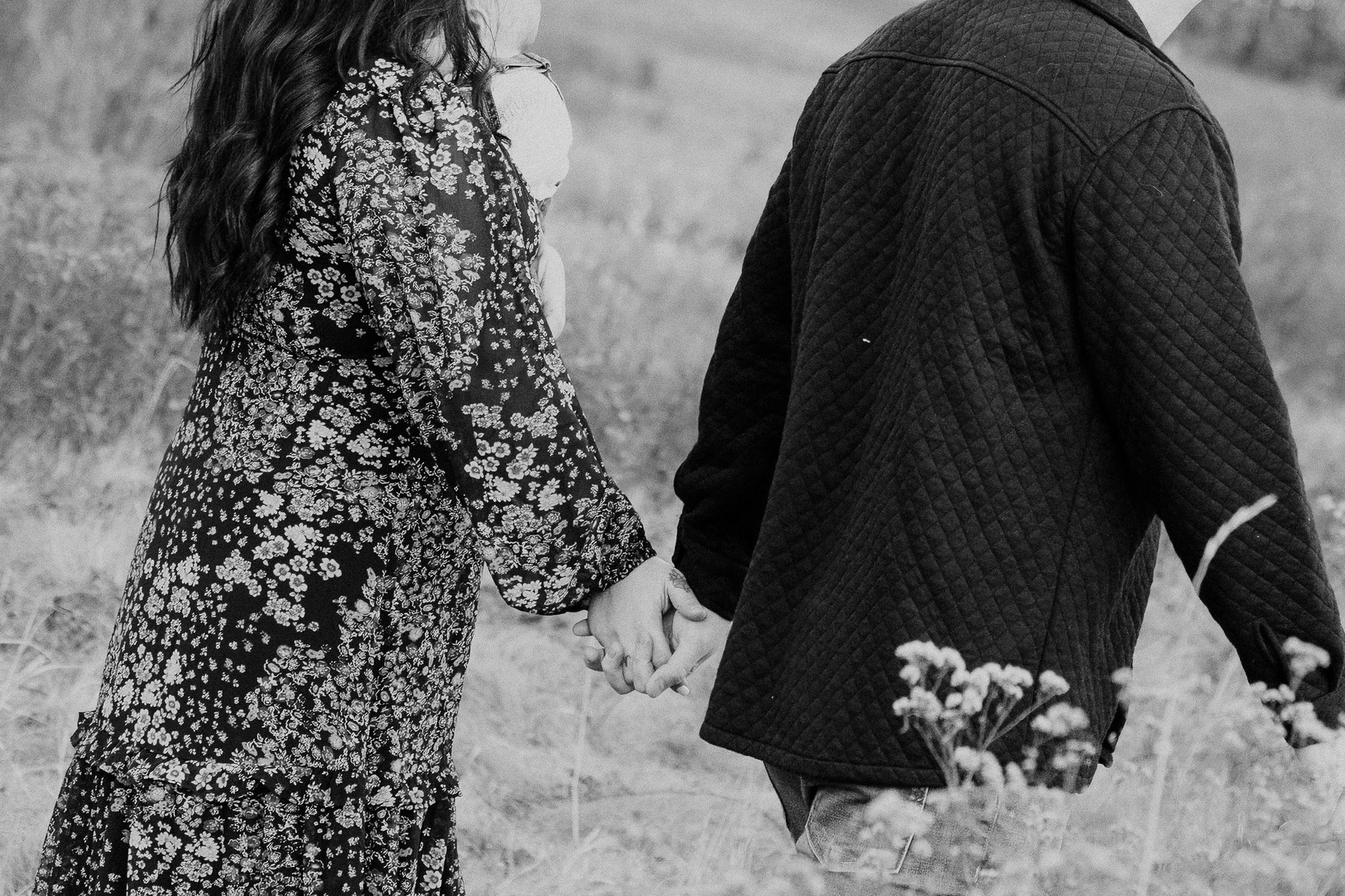 A close-up of a man and woman holding hands in a field, with only their torsos visible, in black and white.