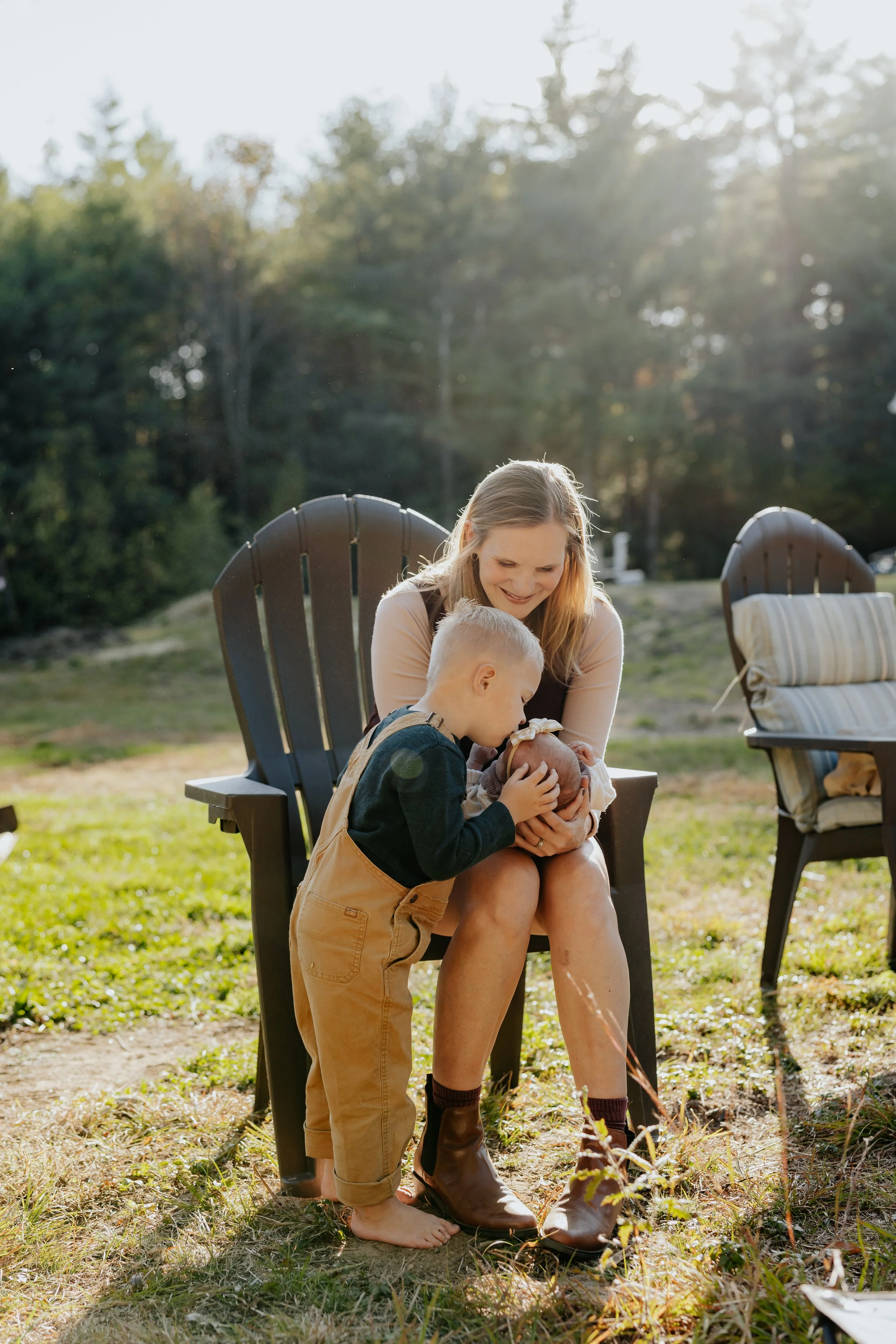 A woman and two children, one kneeling and one sitting, sharing a tender moment with a baby outside on a sunny day, with trees in the background.