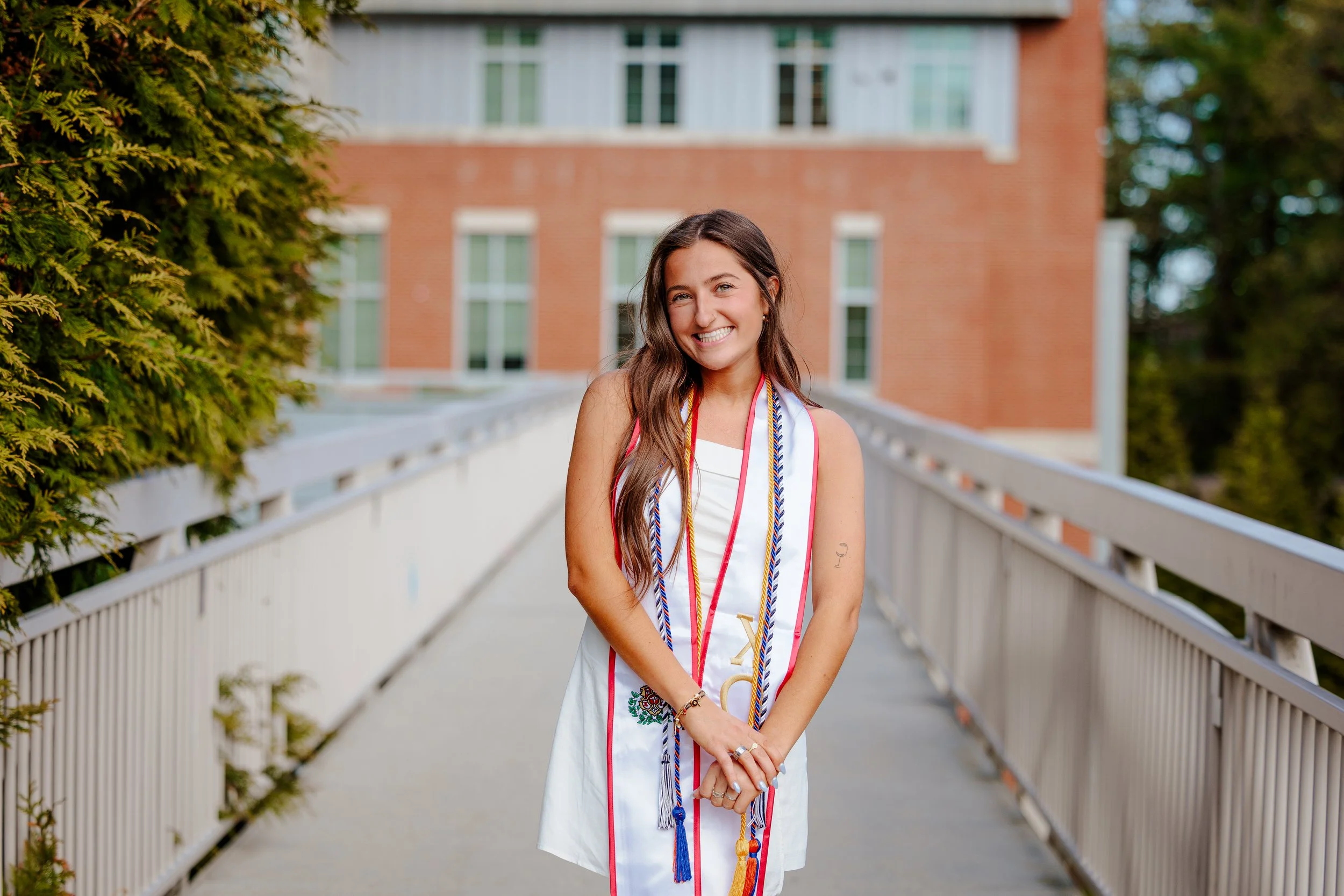 A young woman in a white dress standing on a pedestrian bridge, smiling, with multiple graduation cords around her neck, outdoors in front of a brick building.