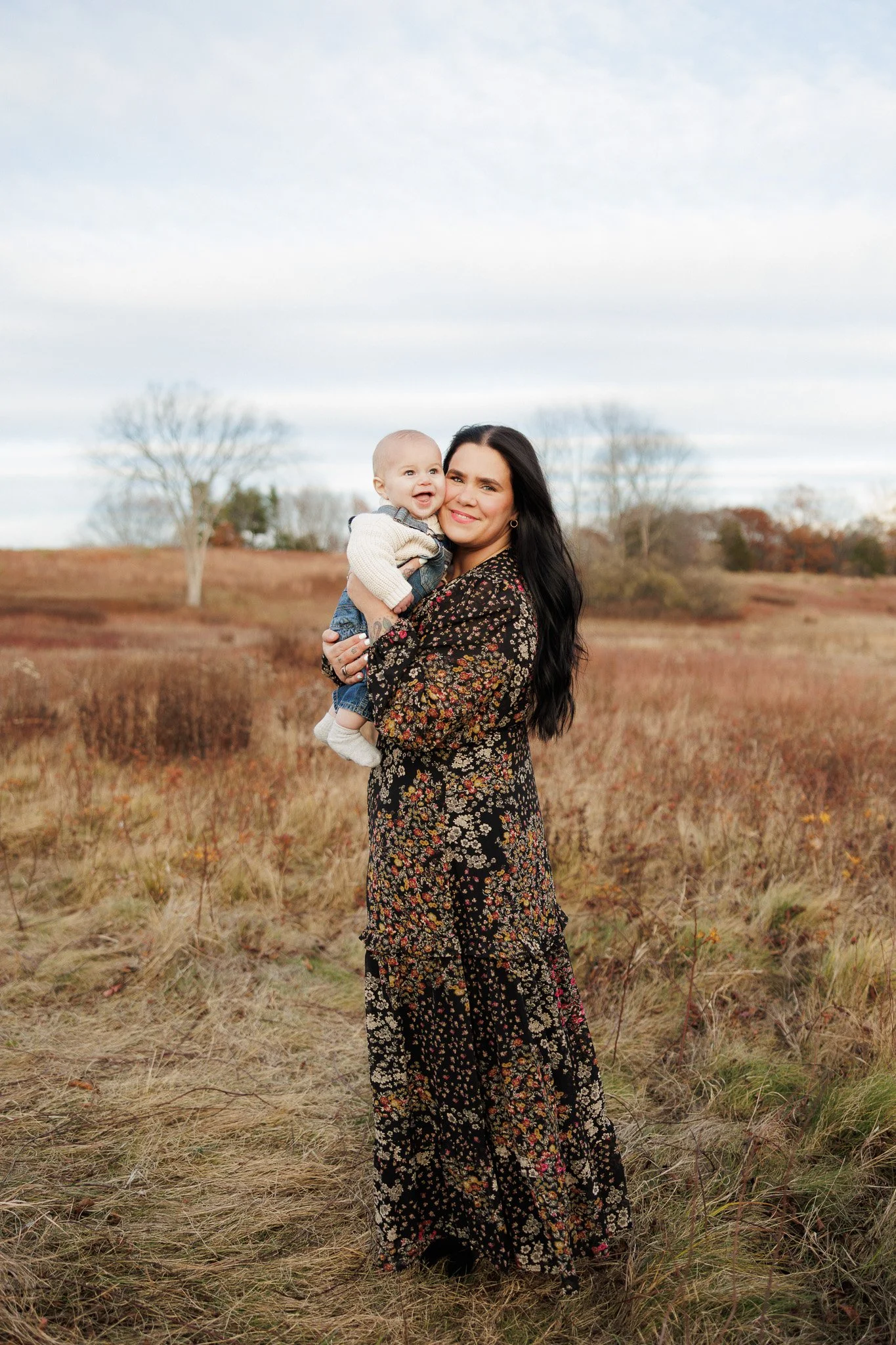 A woman with long black hair wearing a black floral dress holding a smiling baby in an open field with tall grass and trees in the background.