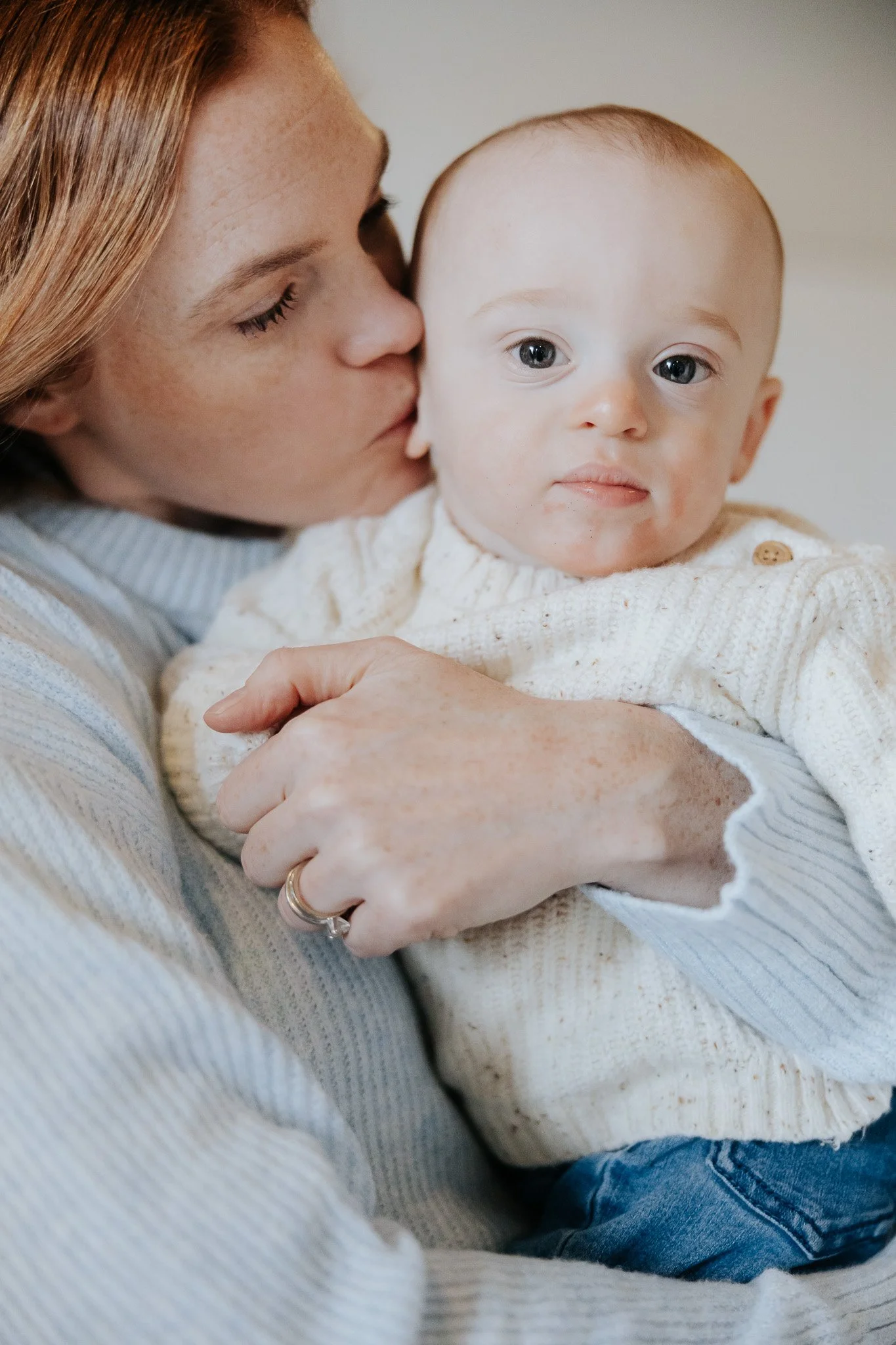 A woman is holding a baby, gently kisses the baby on the cheek, and has her eyes closed. The baby has blue eyes, a soft expression, and is wearing a cream-colored sweater.