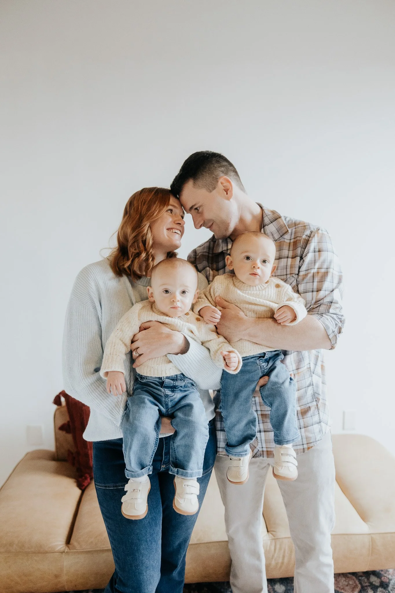 A family of four, including a woman, a man, and two babies, smiling and hugging inside a home, with a beige couch in the background.