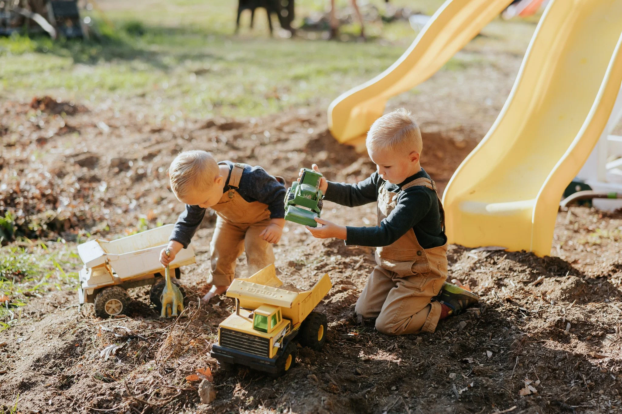 Two young boys playing in a dirt area with toy trucks, near a yellow slide, outdoors on a sunny day.