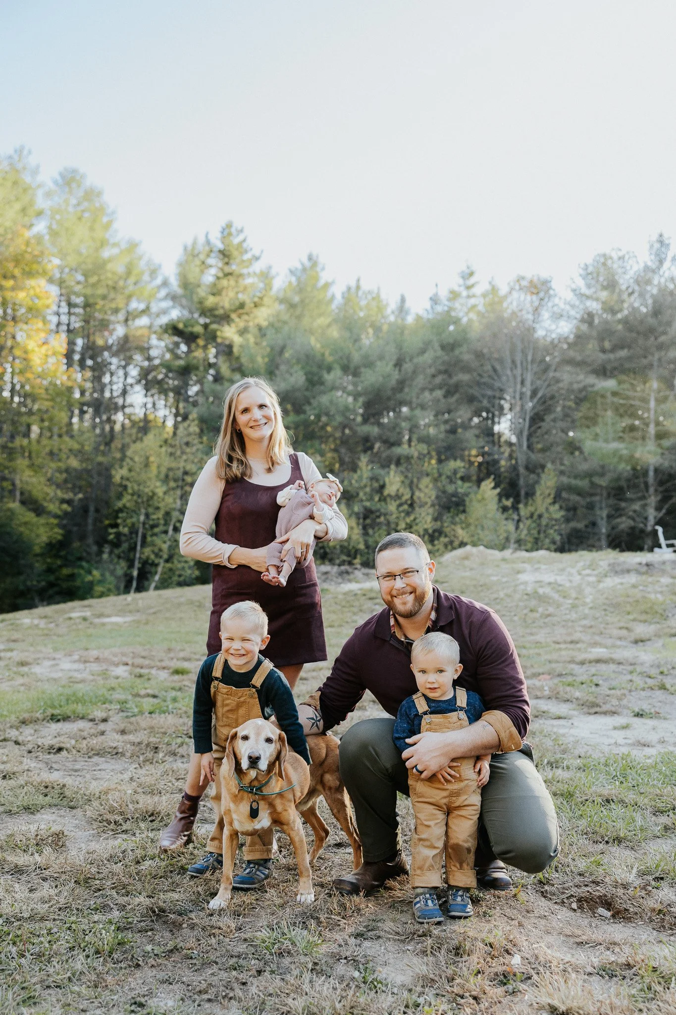 Family outdoor portrait with two children and two dogs in a grassy area with trees in the background.