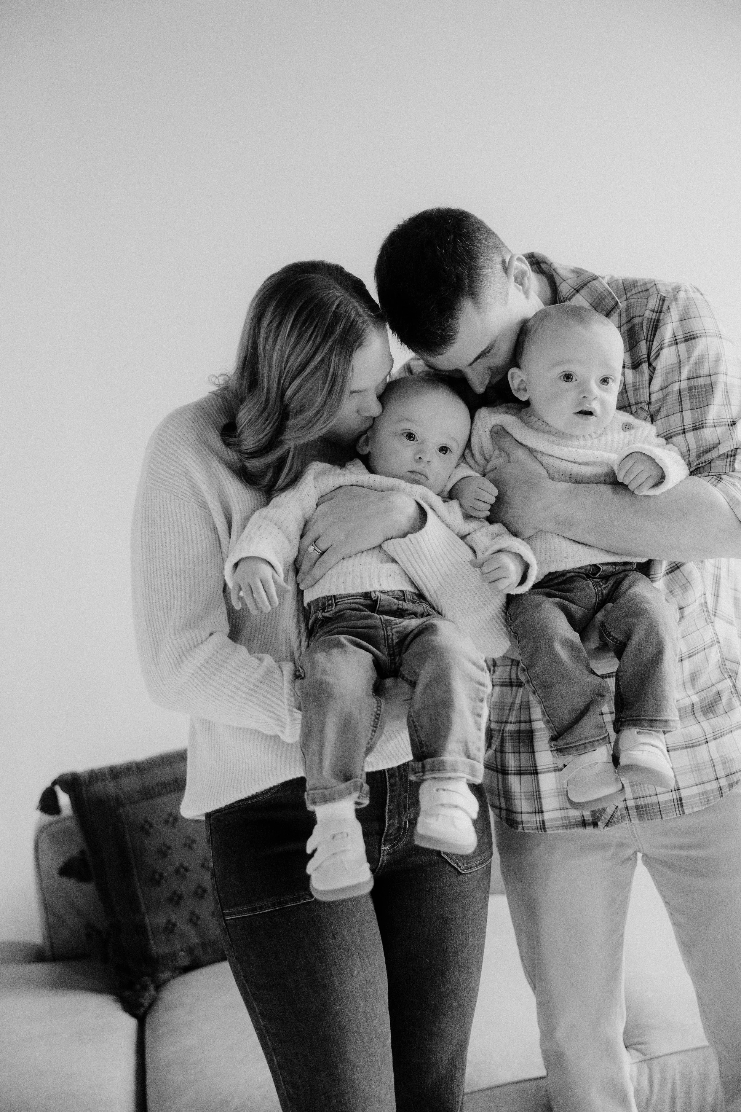 A black and white photo of a family with two adults and two infants. The adults are holding the babies close while standing indoors, with a suitcase in the background.