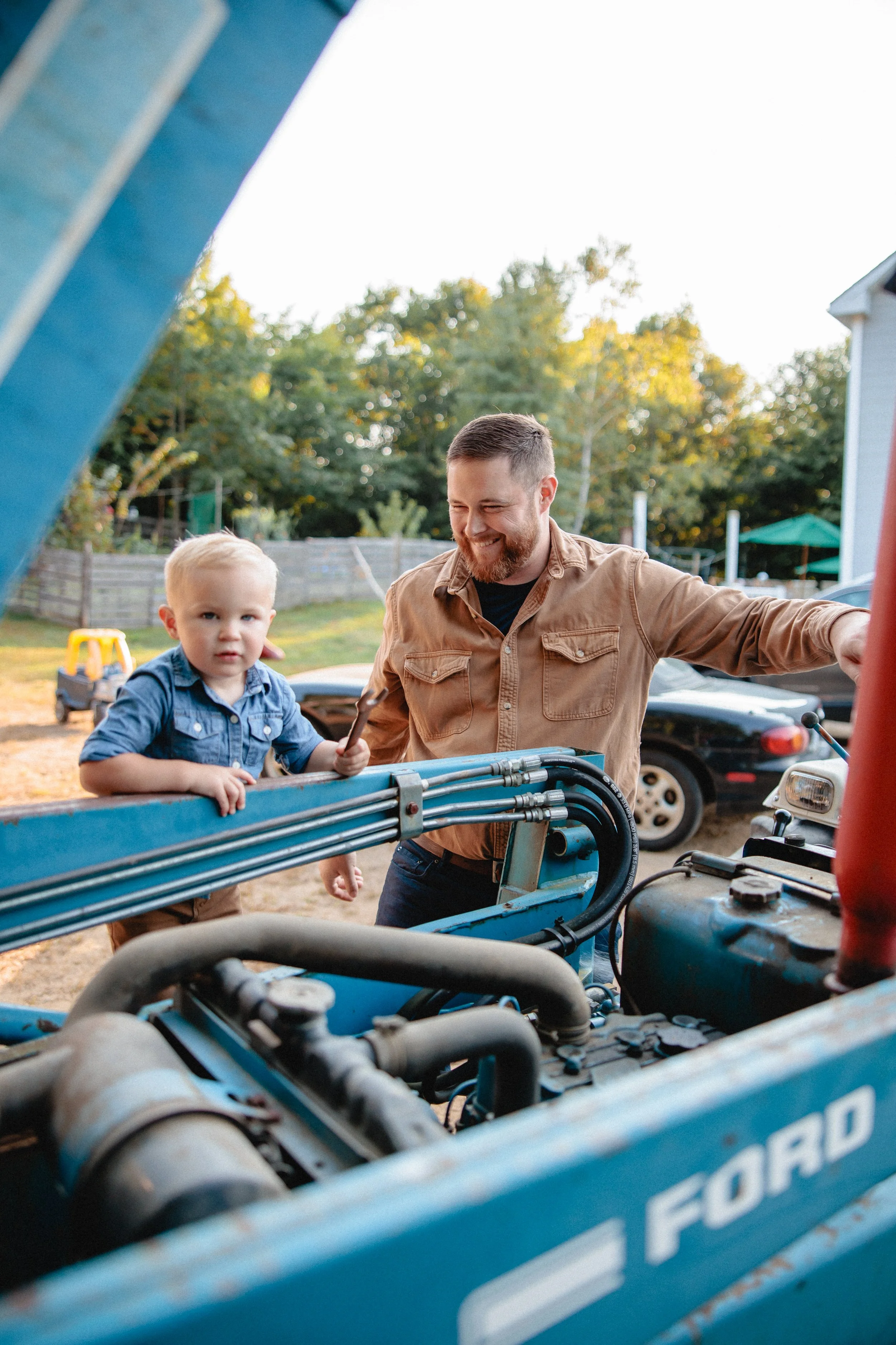 A man and young boy working on a blue tractor outdoors, with cars and trees in the background, during daytime.
