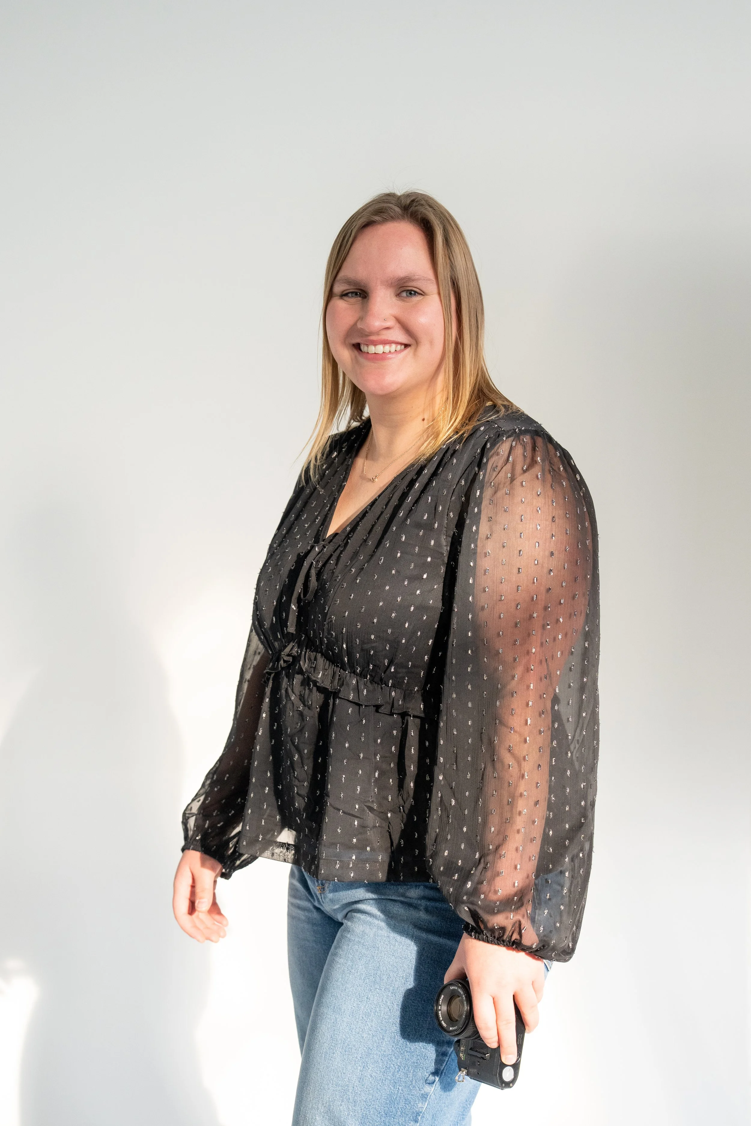 A smiling woman with shoulder-length blonde hair wearing a semi-sheer black blouse with a dotted pattern and jeans, holding a small camera in her right hand, standing against a plain white background.