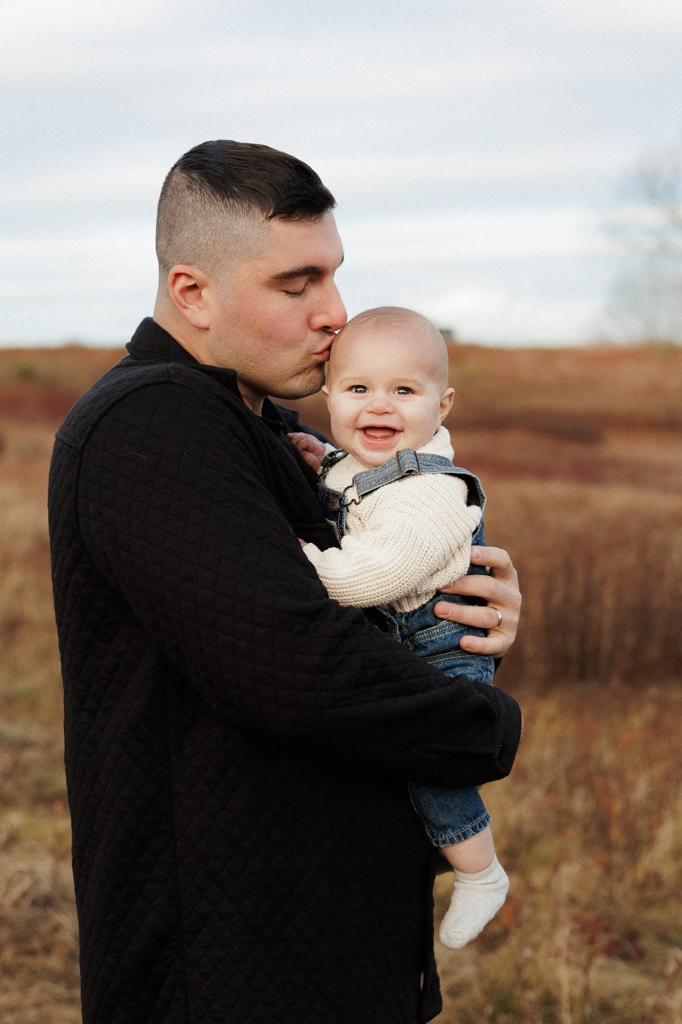 A man kissing a smiling baby girl in an outdoor field.