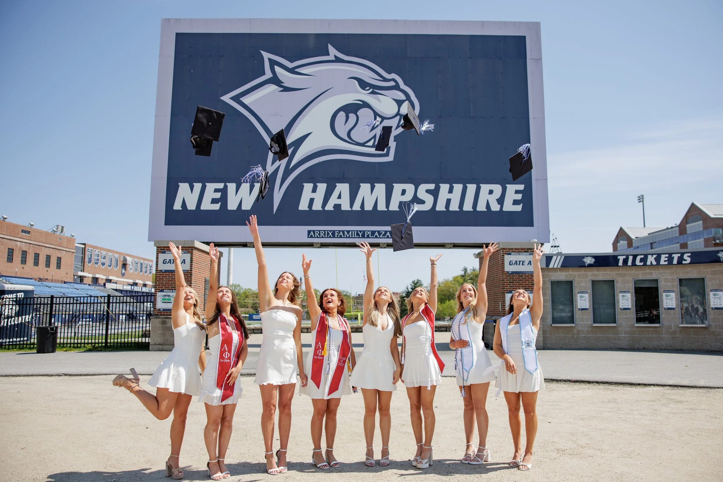 A group of women celebrating graduation outside at a stadium in front of a large New Hampshire sign, with their caps thrown in the air.