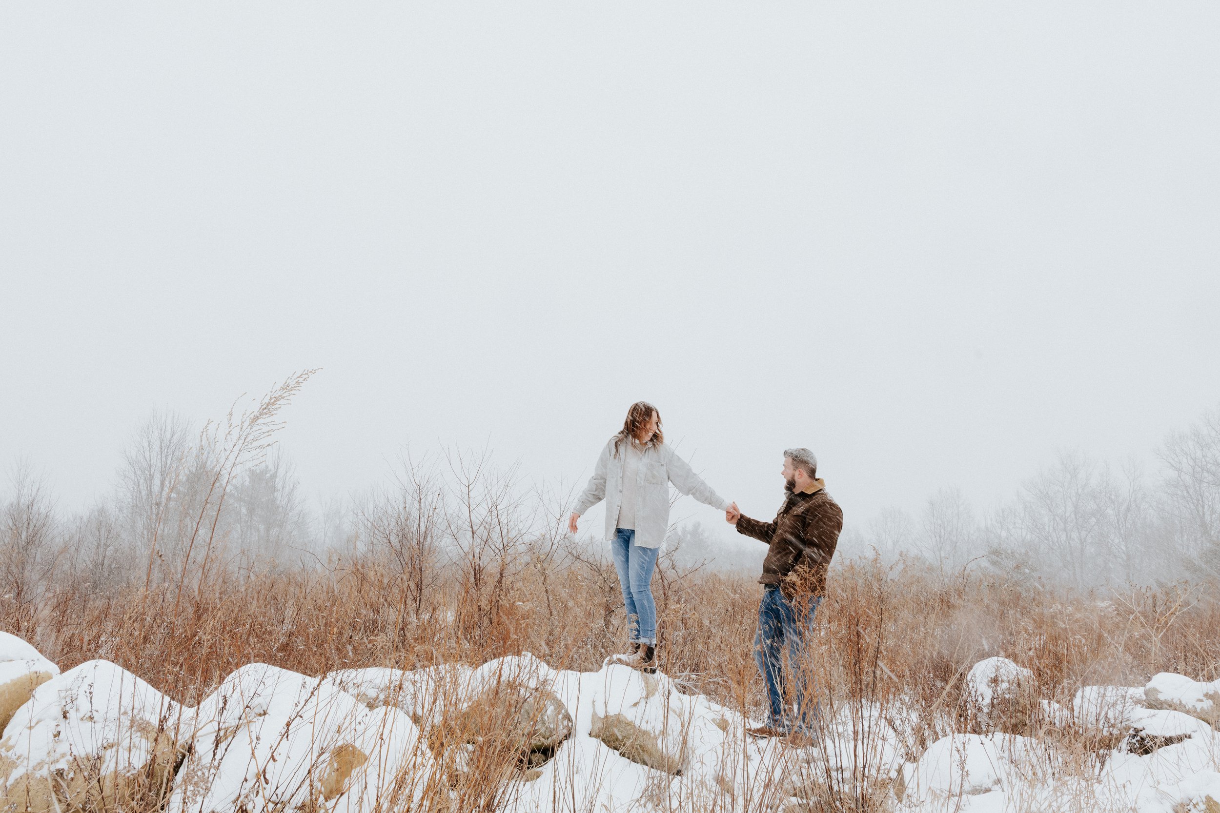A man and a woman holding hands in a snowy field with dry grass and bare trees, overcast sky.