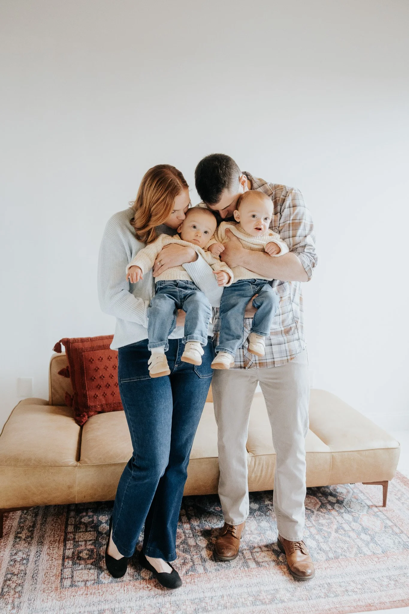 A family of four with two parents and two young children, holding the children and standing in a living room with a beige sofa and a tufted red pillow.