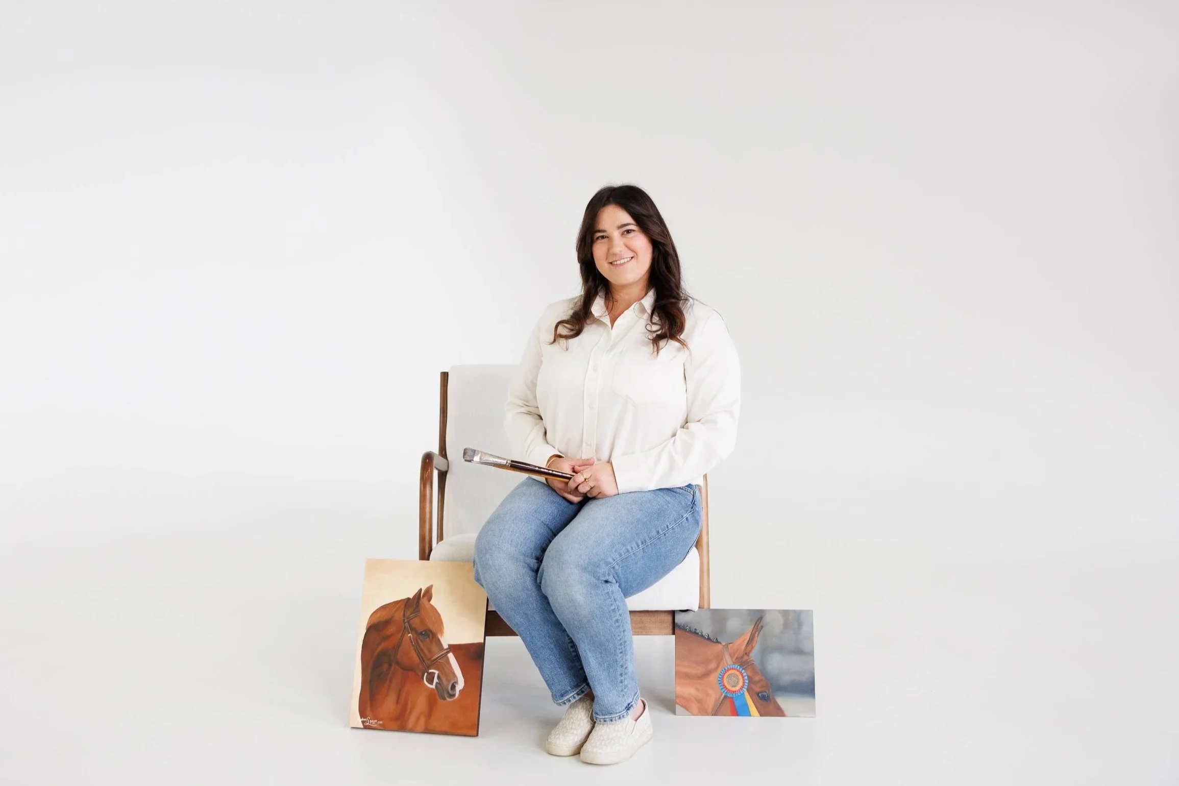 A woman sitting on a white chair, holding a paintbrush, with two horse paintings on the floor beside her, in a white studio background.