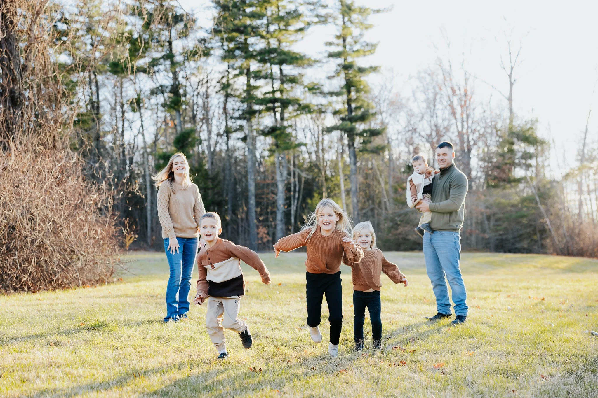 A family of seven enjoying outdoors in a sunlit grassy area, with trees in the background, some children running and two adults holding young children.