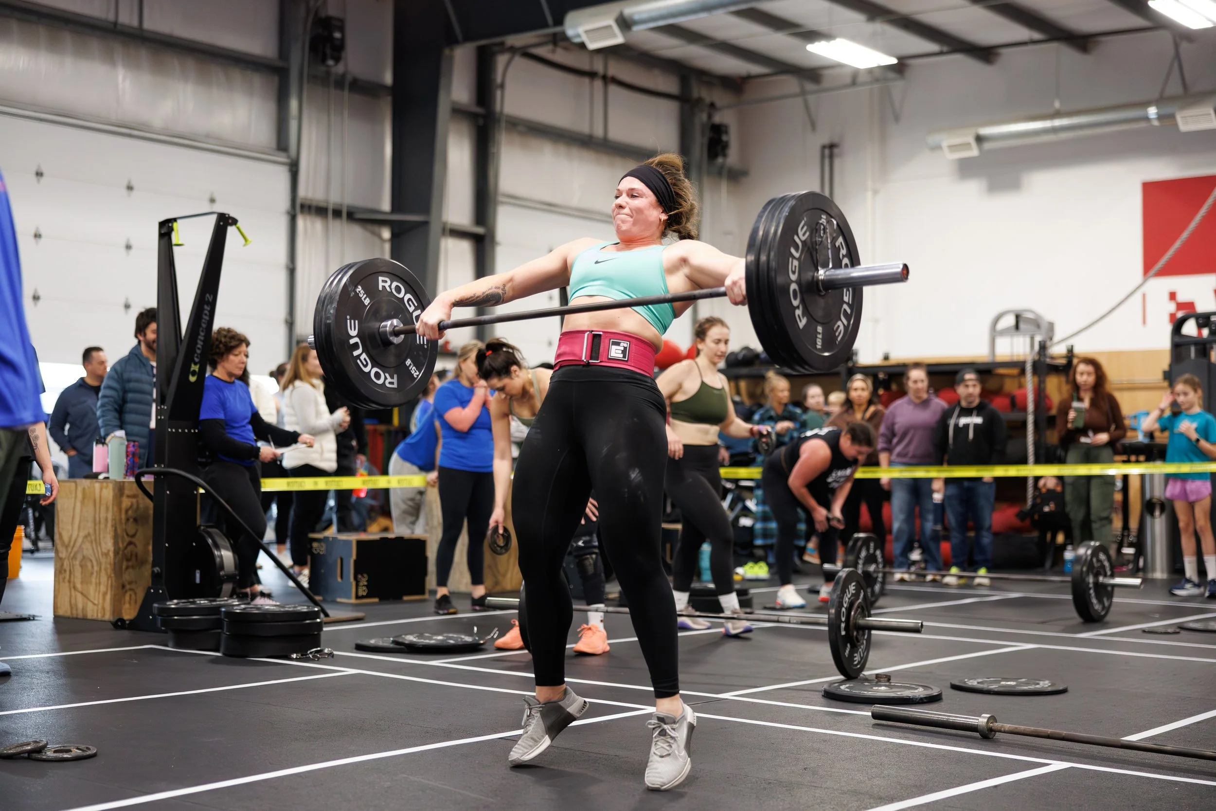 A woman lifting a barbell in a gym during a CrossFit competition, with spectators watching in the background.