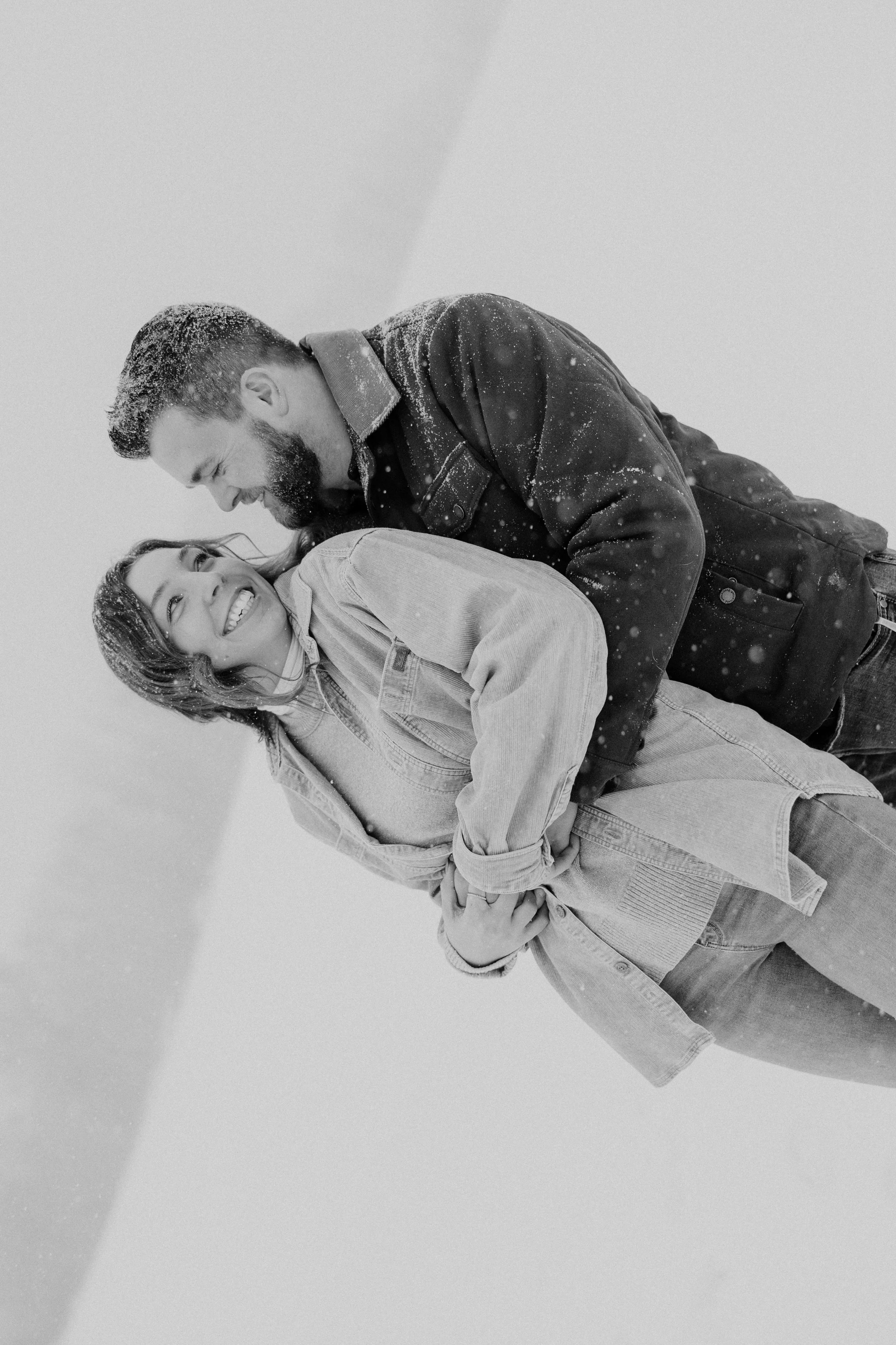 A black-and-white photo of a smiling woman in a denim jacket and a smiling man in a dark jacket, leaning close together indoors on a snowy day.