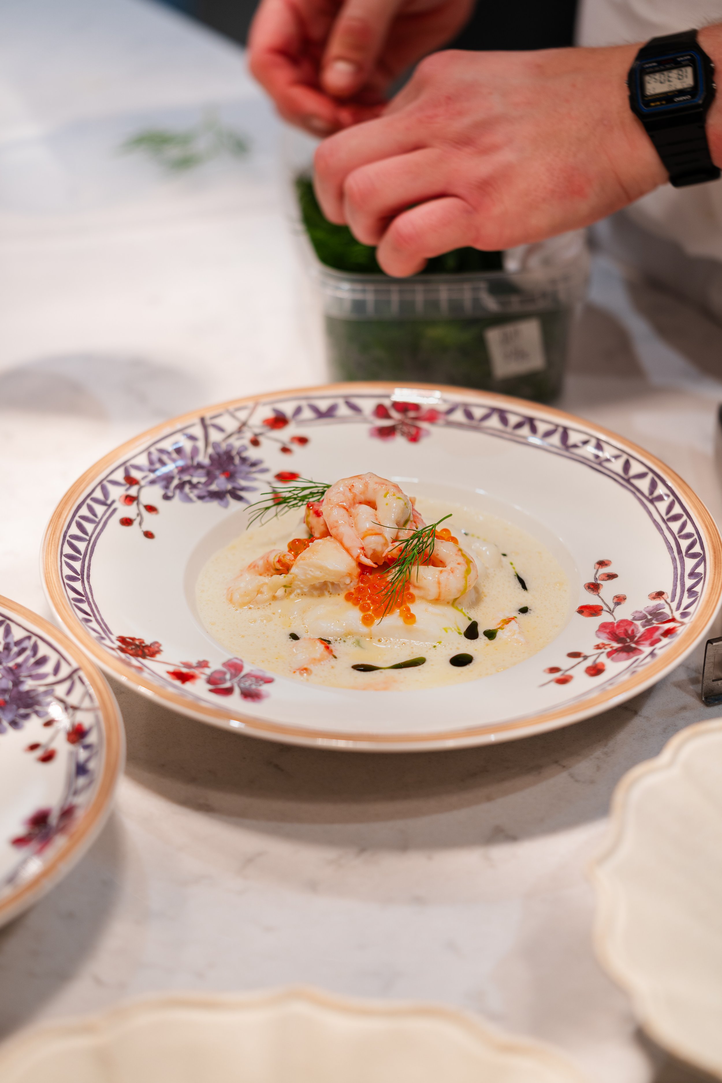 A person prepares a seafood dish with shrimp and red roe garnished with dill, served in a decorated bowl.