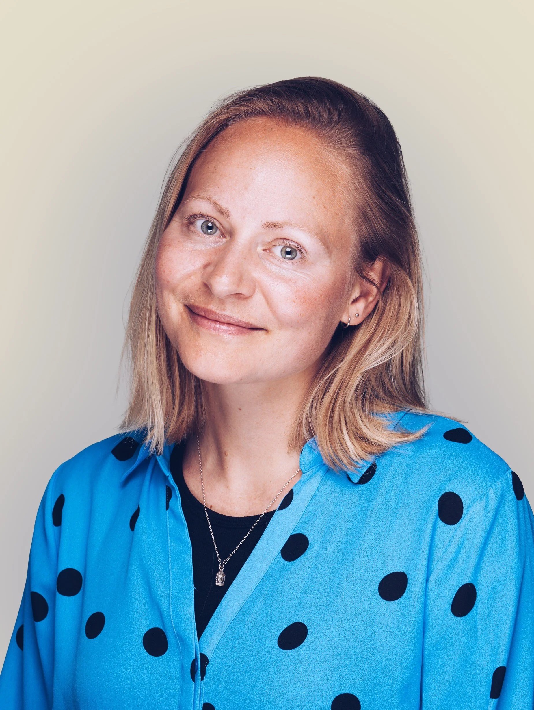 Close-up portrait of a smiling woman with blonde hair and blue eyes, wearing a blue polka-dot blouse and a silver necklace, against a plain light background.