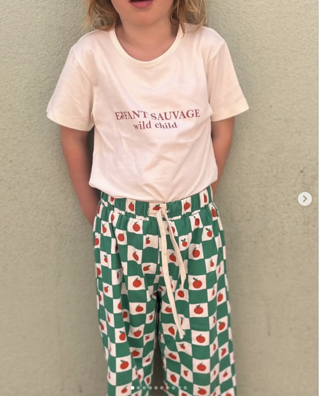A young girl with blonde curly hair wearing a white t-shirt that reads 'ENFANT SAUVAGE wild child' and green and white checkered pants with a tomato pattern, standing against a neutral wall.