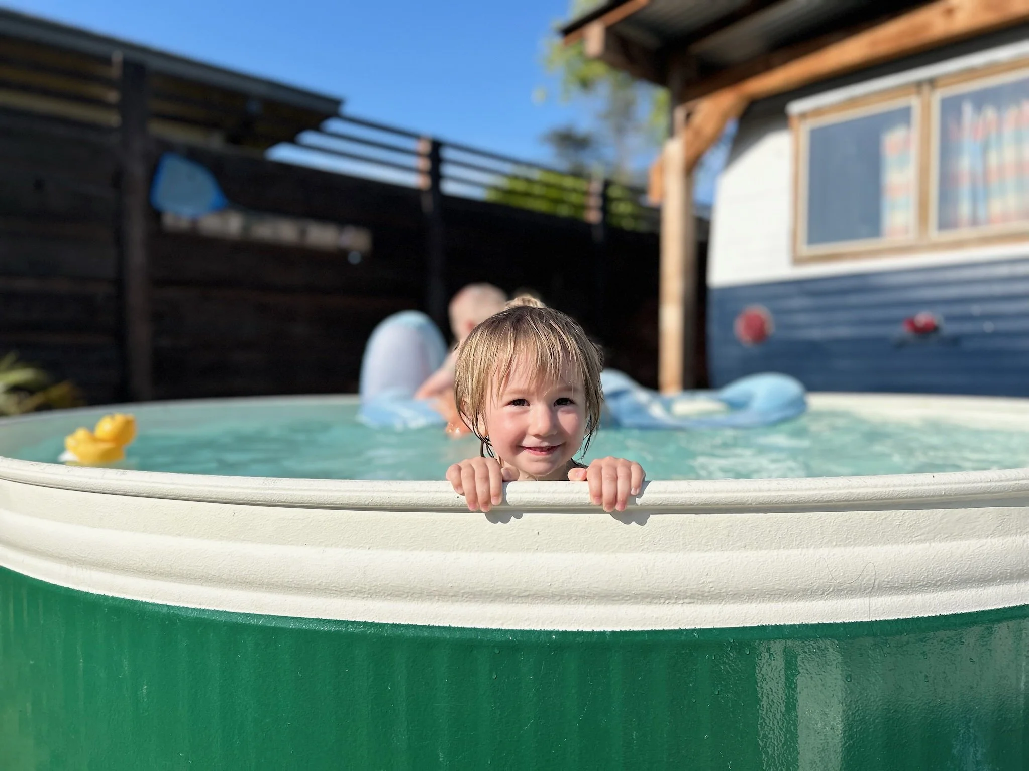 A young child with wet hair smiling and holding onto the edge of a small green and white inflatable pool, with a duck toy floating in the water, outside in a backyard on a sunny day.