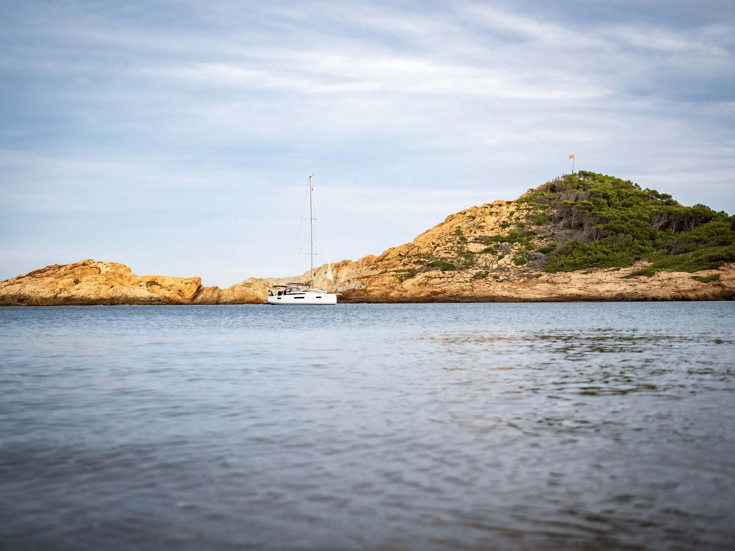 A sailboat floating near rocky islands with green vegetation, under a cloudy sky.