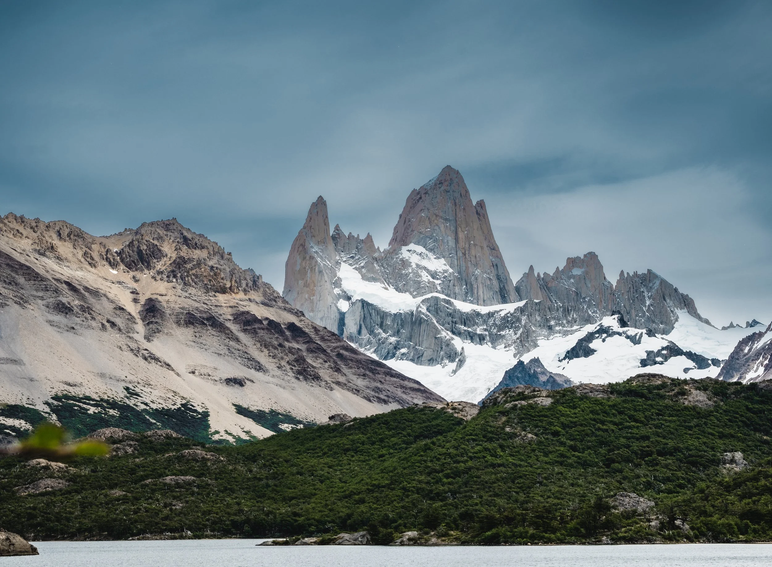 Snow-capped mountain peaks with a lake and green forest in the foreground, under a cloudy sky.