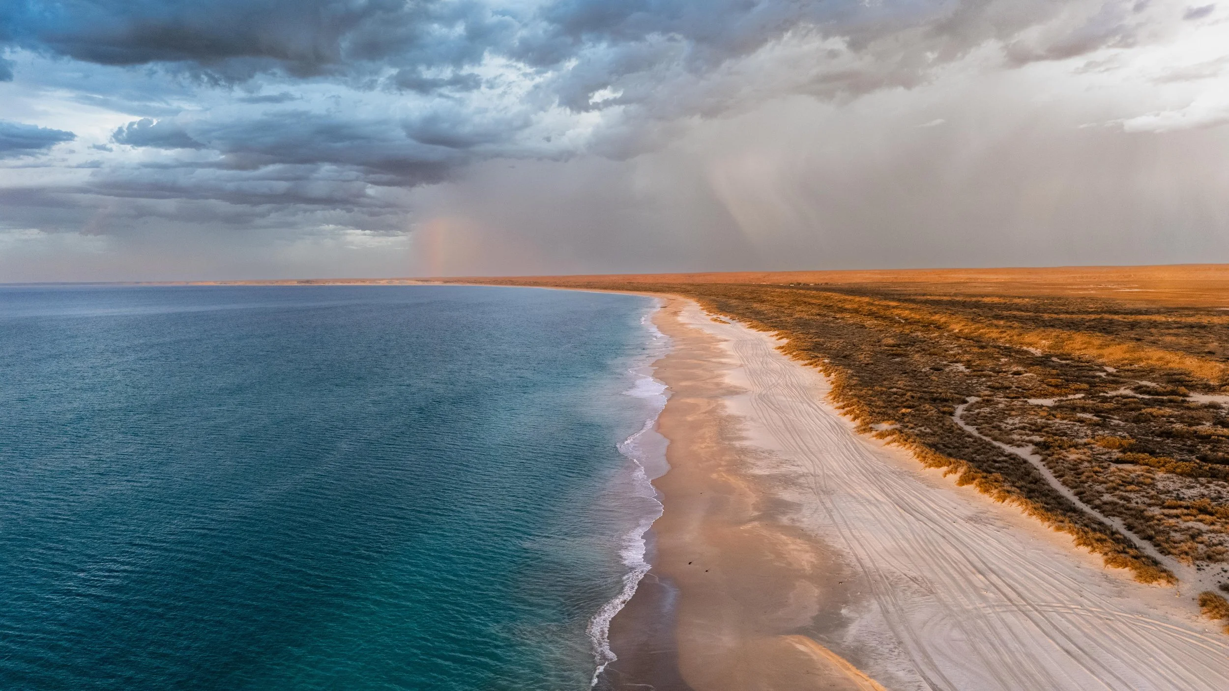 Aerial view of a coastline with blue ocean on the left and sandy beach on the right, with a cloudy sky overhead.
