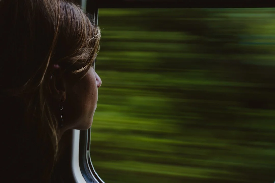 A woman with brown hair looking out of a bus window at green blurred scenery.