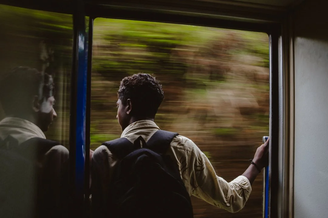 A person with a backpack looking out of a moving train window, with trees passing by quickly.
