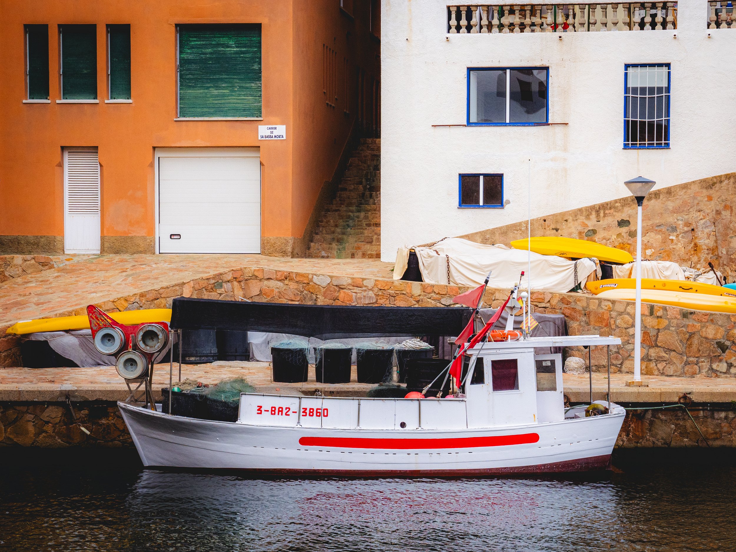 A small white boat with a red stripe, moored along a stone dock, with two yellow kayaks and installations on the dock, beside colorful residential buildings.