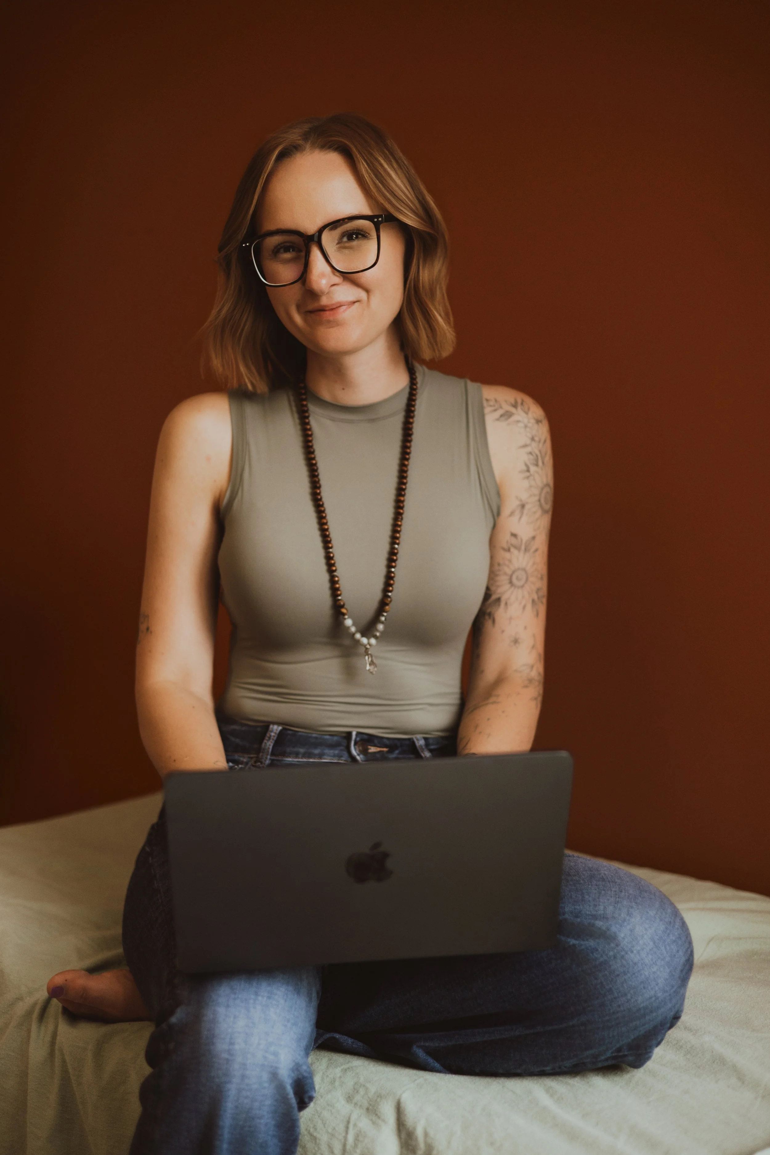 A woman with glasses and tattoos sitting on a bed, using a laptop, with a battery and phone charging cord in the foreground.
