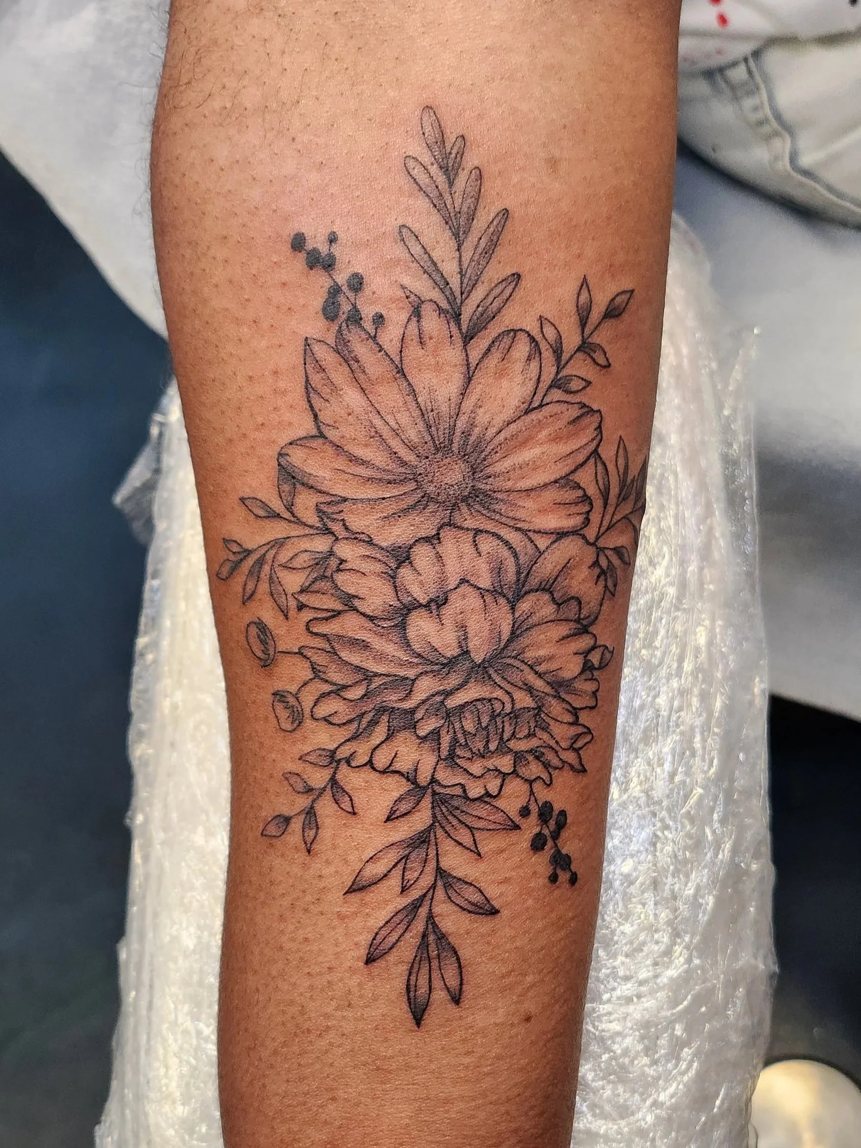 Line drawing tattoo of a bouquet of flowers with various leaves on a person's arm.